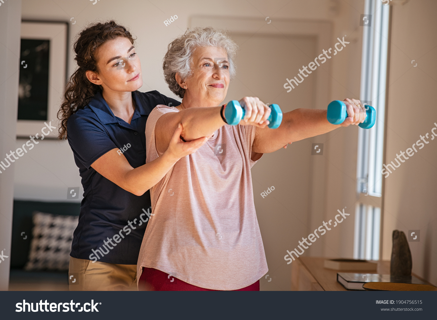 Old woman training with physiotherapist using dumbbells at home. Therapist assisting senior woman with exercises in nursing home. Elderly patient using dumbbells with outstretched arms. #1904756515