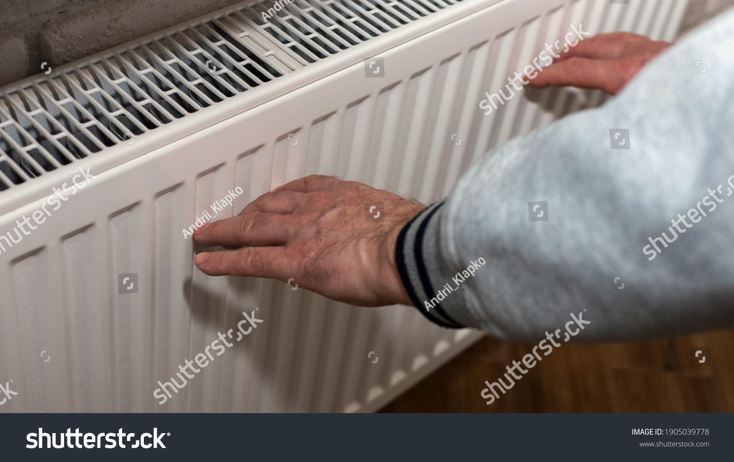 An elderly man warms his hands near a heating radiator, a man froze, a man warms himself near a fireplace. A man warms himself up by an electric battery #1905039778