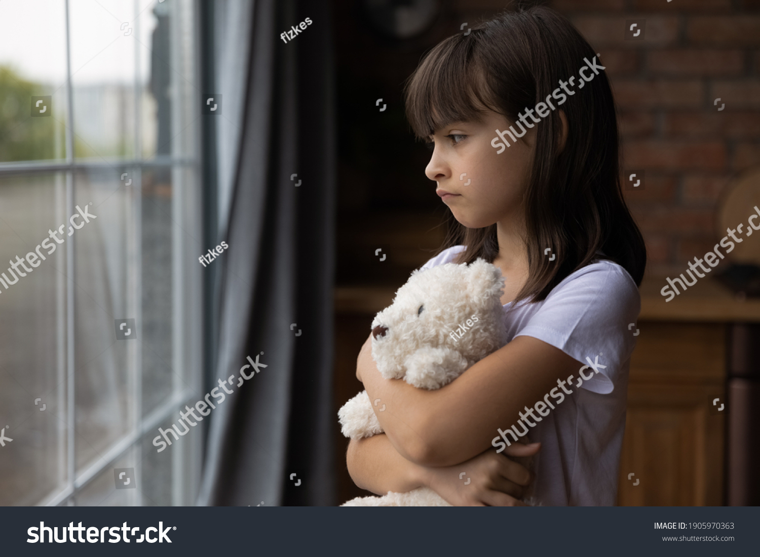 Close up lonely little girl hugging toy, looking out window, standing at home alone, upset unhappy child waiting for parents, thinking about problems, bad relationship in family, psychological trauma #1905970363