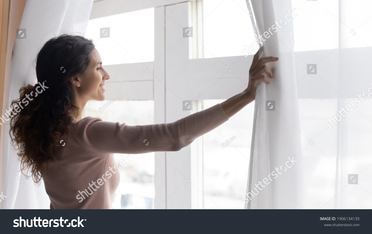 Smiling young dreamy woman opening curtains, looking outside window in morning. Sincere millennial caucasian lady breathing fresh air, starting new day or enjoying peaceful weekend alone at home. #1906134139