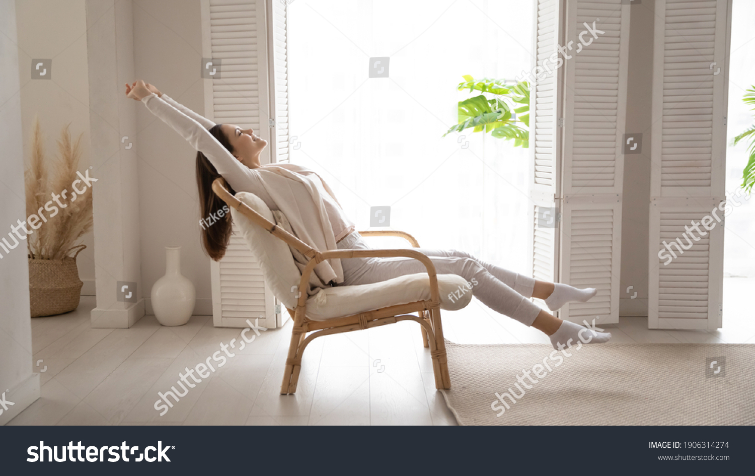Happy millennial Caucasian woman sit in cozy chair in design home stretch relax breathe fresh air. Smiling young female renter or tenant rest in armchair relieve negative emotions enjoying weekend. #1906314274