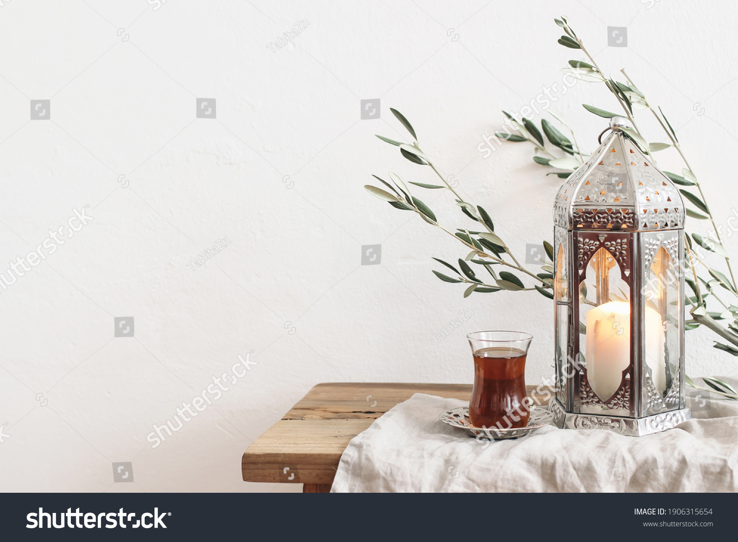 Ramadan Kareem greeting card, invitation. Silver lantern with burning candle. Turkish tea in glass on saucer. Green olive tree branches on old wooden table background. Muslim Iftar dinner. Front view. #1906315654