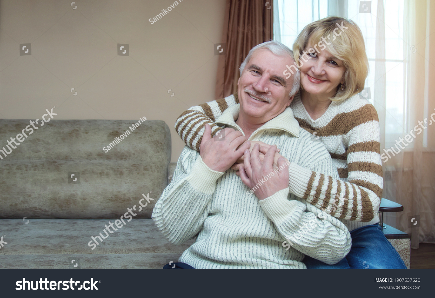 A married couple of 50-60 years old are hugging while sitting on the sofa at home. Looking into the camera. The concept of a long happy marriage. Place for text. #1907537620