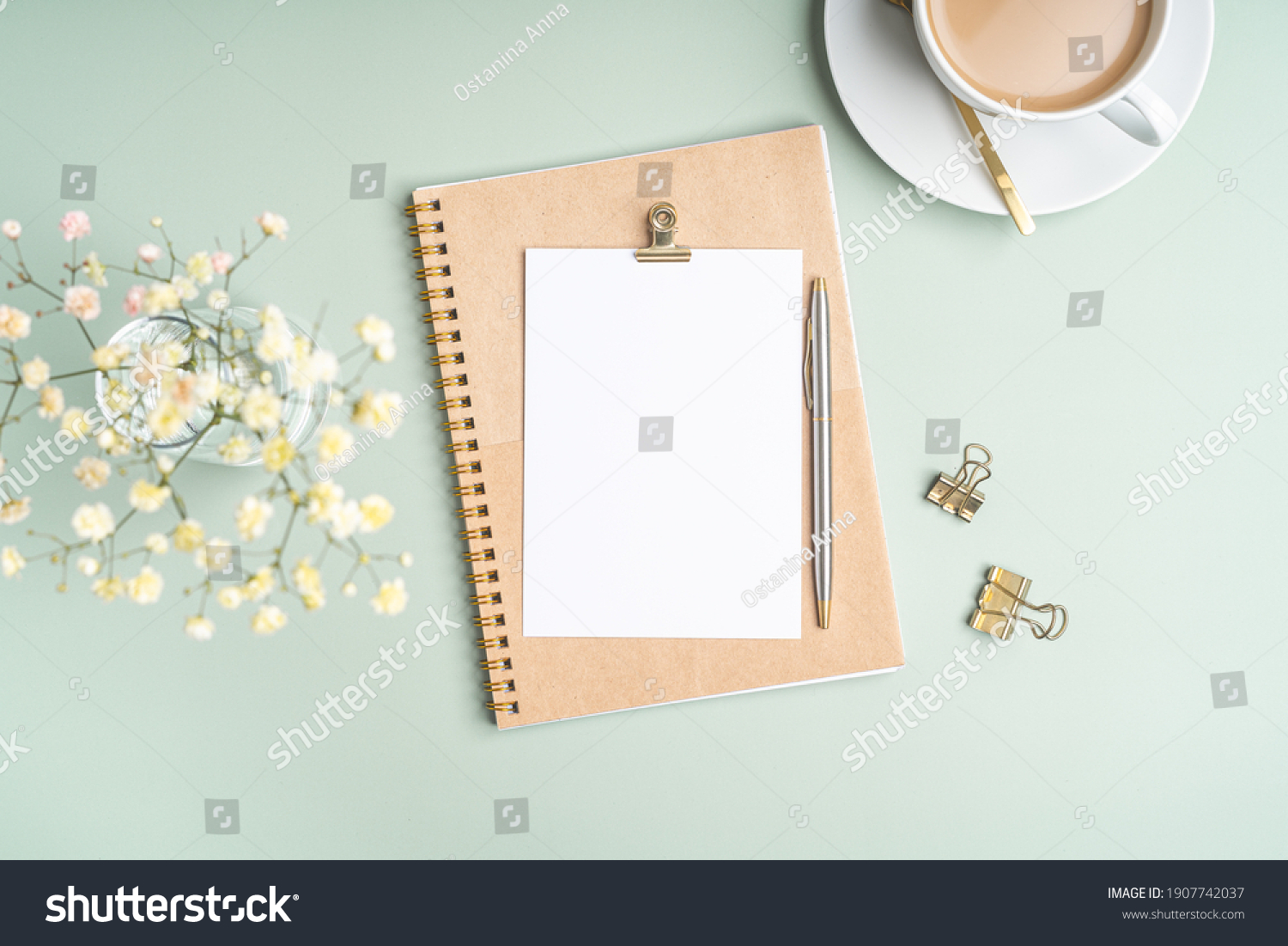 Top view blank paper Notebook, flowers, golden paper binder clips, cup of coffee and pen. Desktop mock up, Flat lay of green working table background with office equipment, mockup greeting card #1907742037