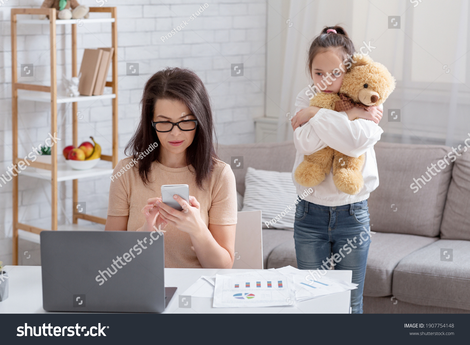Lonely teen girl hugging teddy bear while her busy mom working online from home, not paying attention to child. Depressed kid feeling neglected, missing her parent. Covid-19 quarantine family problems #1907754148