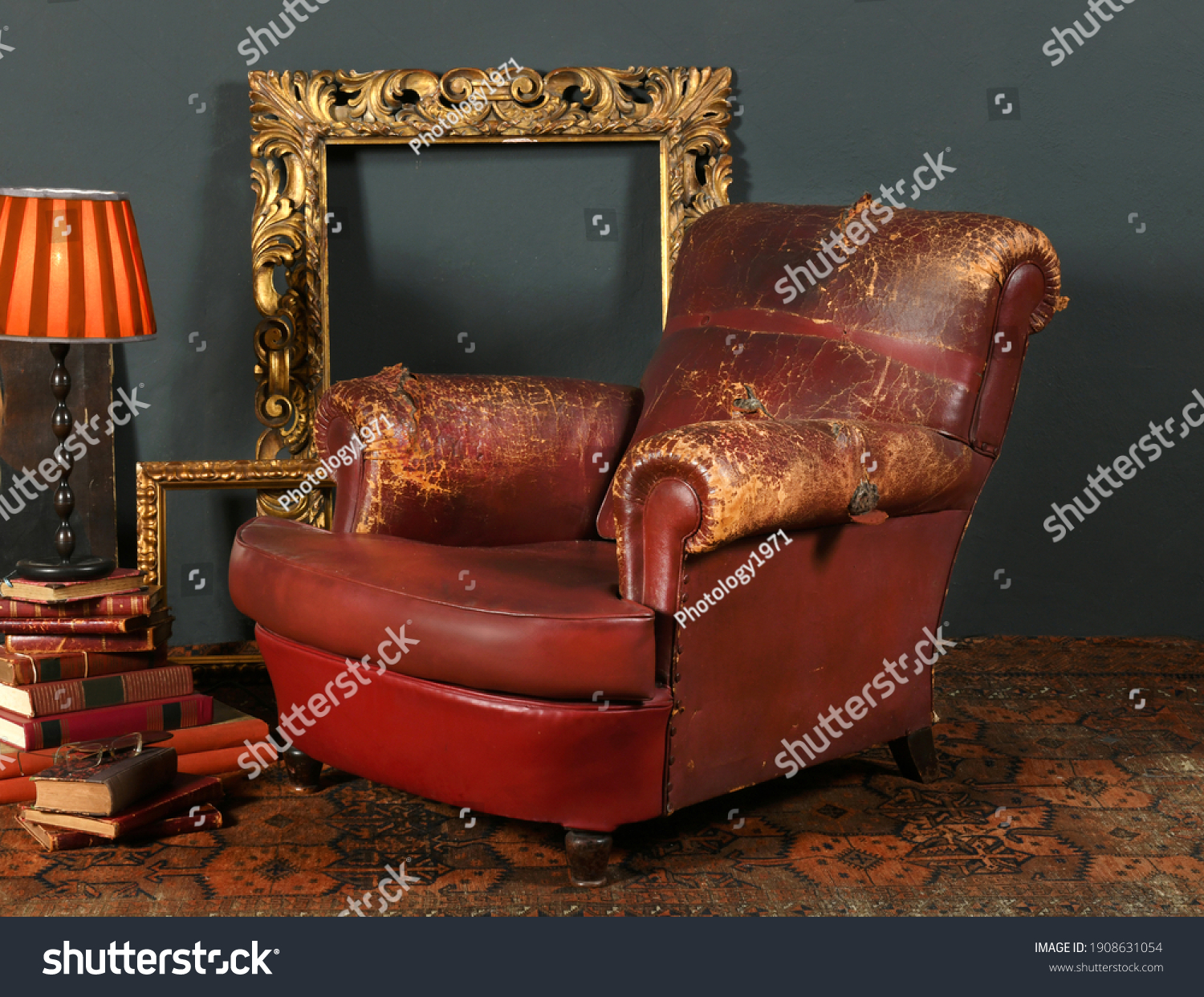 Old shabby red leather armchair placed near vintage decorative frames and lamp in room with retro style interior #1908631054