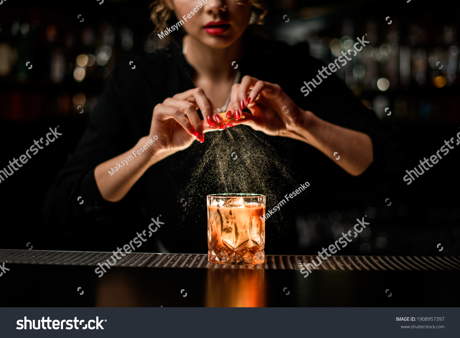 female bartender gently sprinkles orange peel juice on glass with cold alcoholic cocktail on the bar counter #1908957397