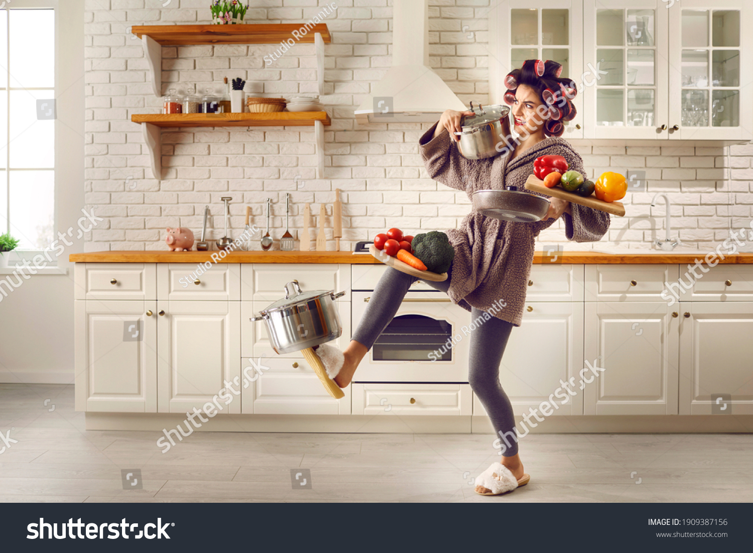 Tired housewife cooking food and carrying lots of stuff. Frail slim young woman making meal at home, holding multiple heavy cooking pots and kitchen saucepans, balancing cutting boards with vegetables #1909387156