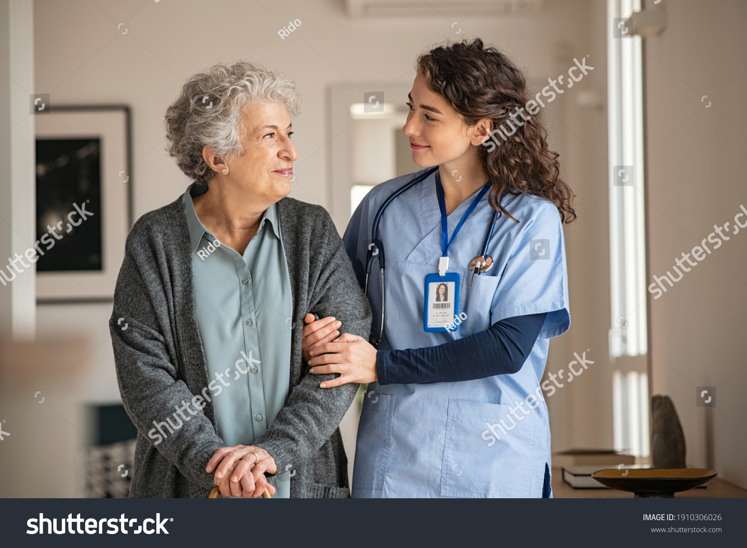 Young caregiver helping senior woman walking. Nurse assisting her old woman patient at nursing home. Senior woman with walking stick being helped by nurse at home. #1910306026