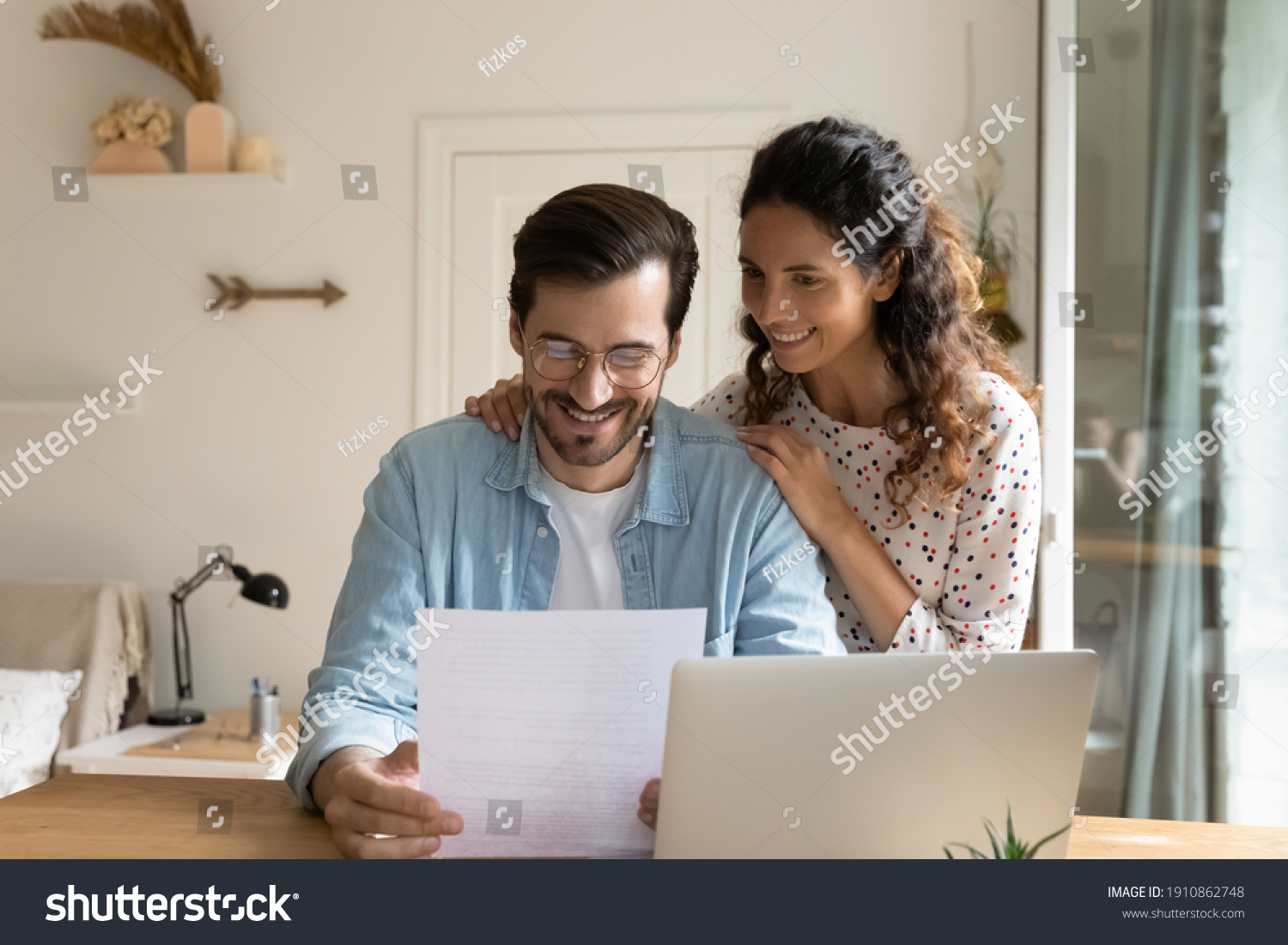 Happy young wife embrace shoulders of beloved husband reading official paper letter of getting job promotion loan mortgage approval. Glad married couple impressed with perfect news received by mail #1910862748