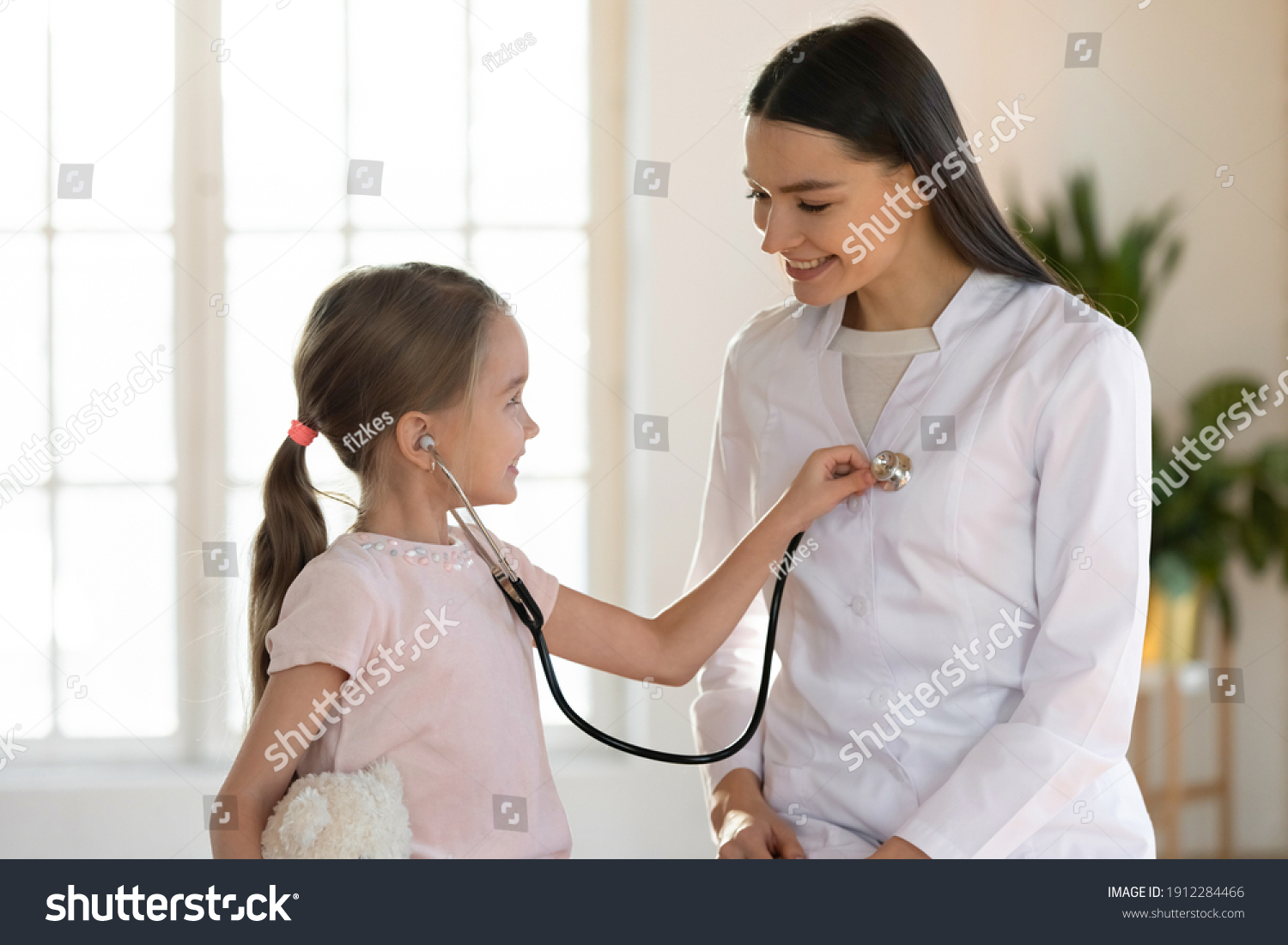Cute small girl child hold stethoscope listen to female nurse heart at consultation in hospital. Caring woman doctor have fun play with little kid patient in clinic. Children healthcare concept. #1912284466