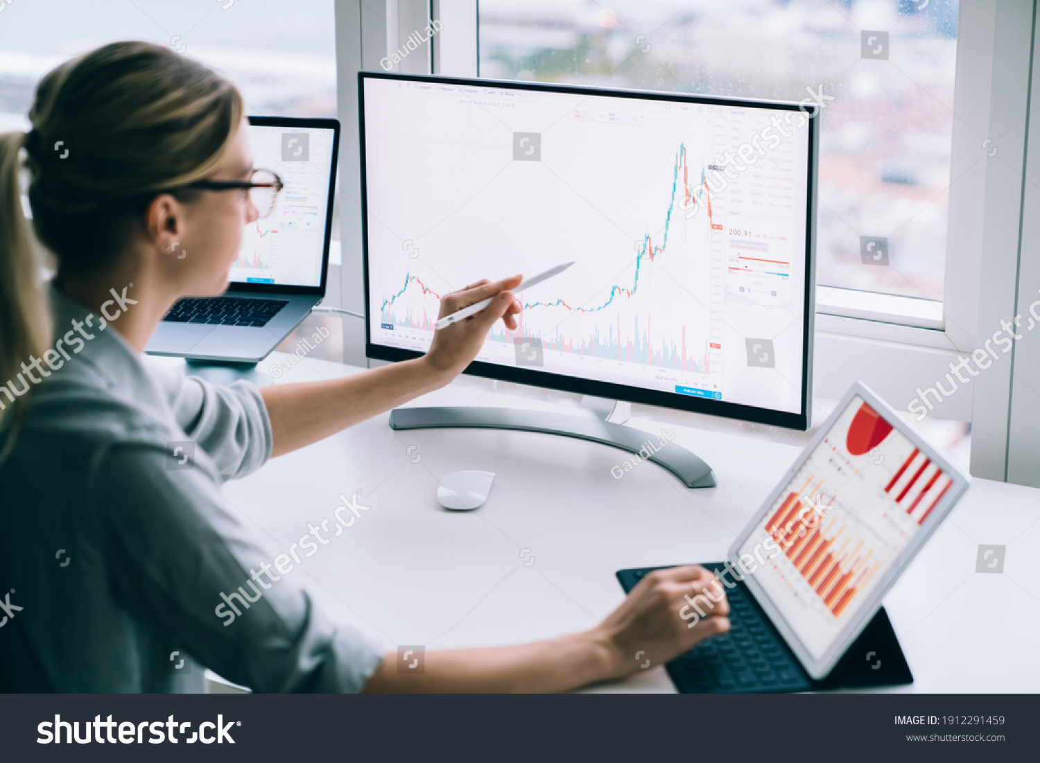 Side view of anonymous young female analyst pointing with stylus at desktop computer while studying chart near tablet at work #1912291459
