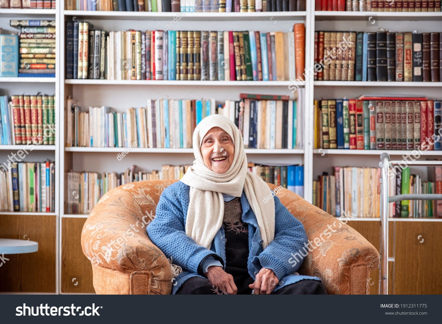 Arabic old woman sitting on couch in her home with a library background #1912311775