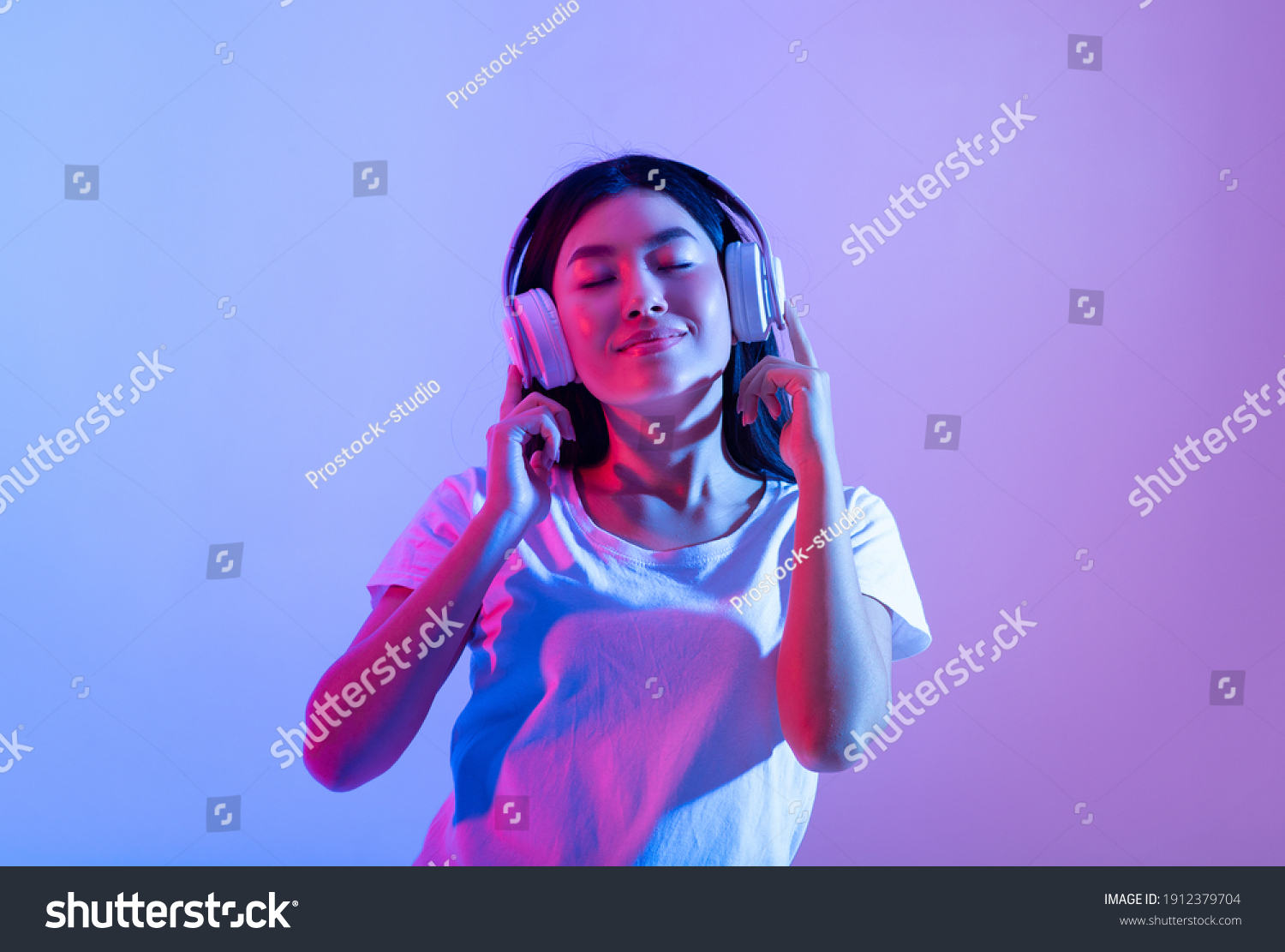 Stay alone at home during covid-19 and relax at spare time with music. Smiling calm millennial asian lady in modern headphones with closed eyes enjoying song in audio app, in neon, studio shot #1912379704