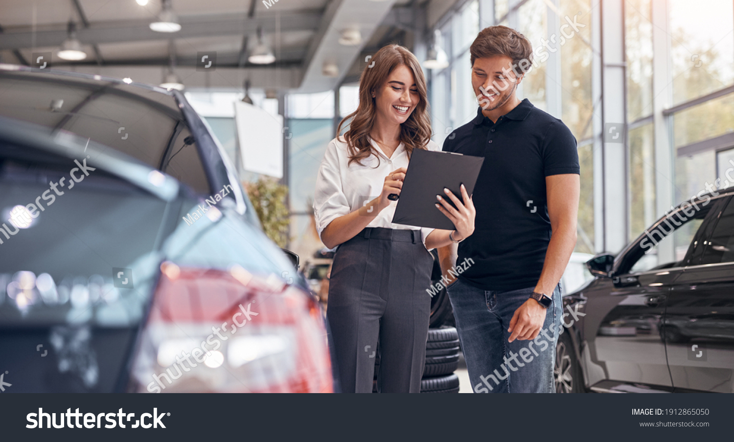 Positive young professional saleswoman demonstrating document to male customer buying new car in dealership #1912865050