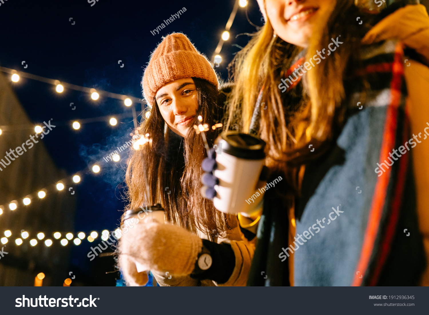 Amazing women friends in gray coat and pink hat walking down the street with sparkler. Adorable females students in winter outfit spending time outdoor and looking at Bengal light with smile. #1912936345