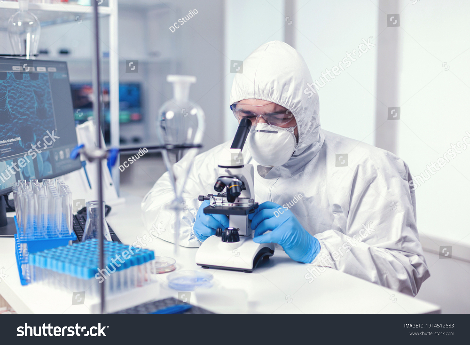 Analyzing virus in microbiology lab using microscope wearing ppe suit and glasses. Virolog in coverall during coronavirus outbreak conducting healthcare scientific analysis. #1914512683