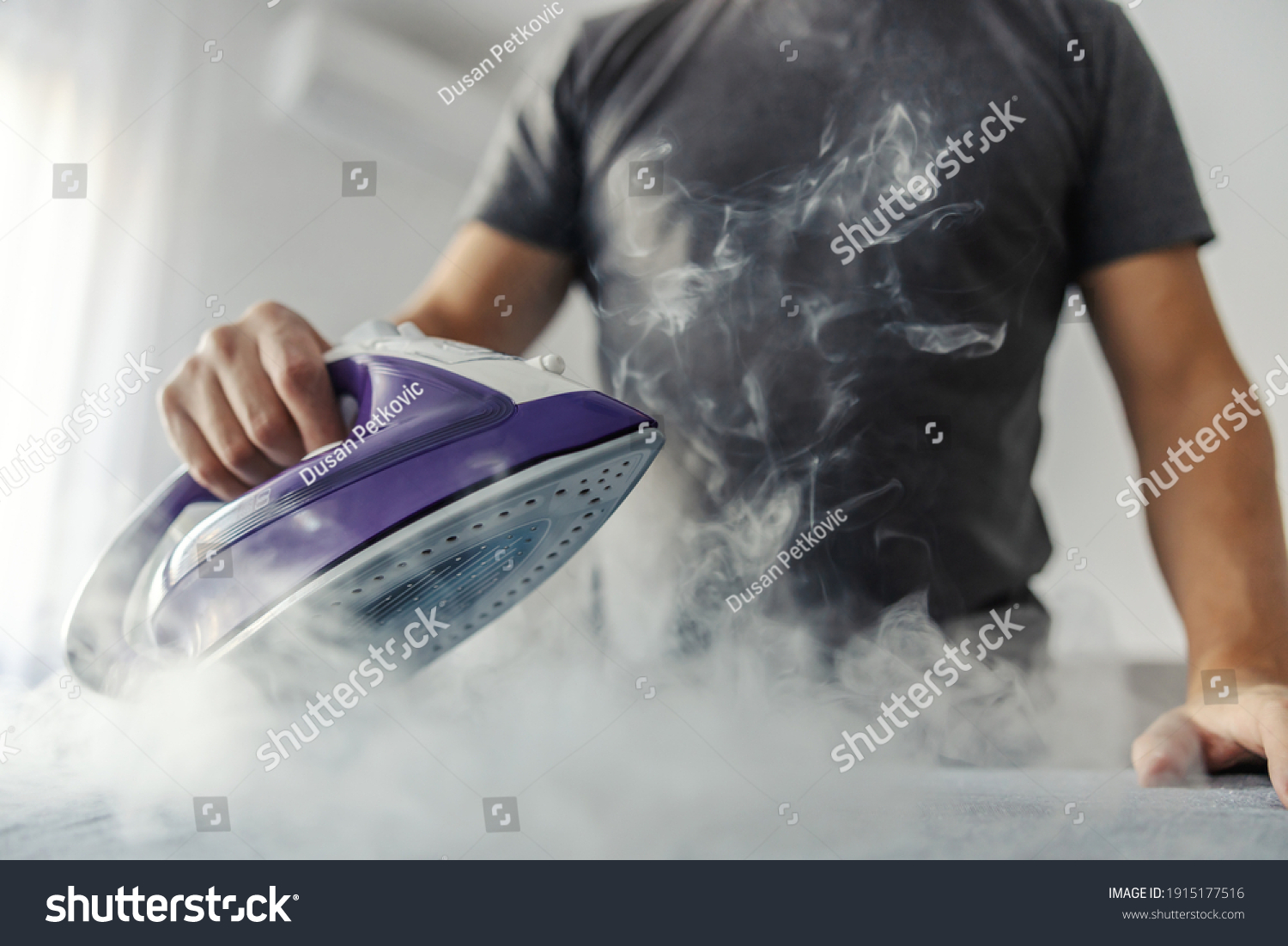 The hot steam from the iron. Powerful film effect of steam on photography. A close-up of a man's body in a grey t-shirt ironing clothes on an ironing board #1915177516