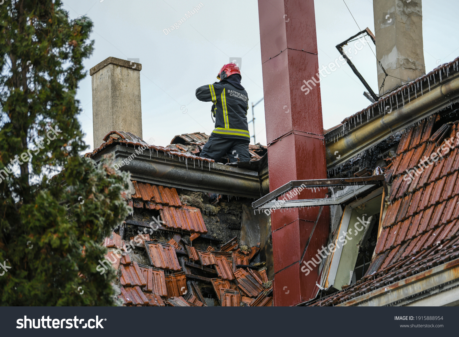 Firefighter inside a collapsed roof of a house is looking for survivors. House rooftop damage. Charred roof trusses and burnt furniture. A blazing fire burns roof rafters. Translation:''Firefighters'' #1915888954
