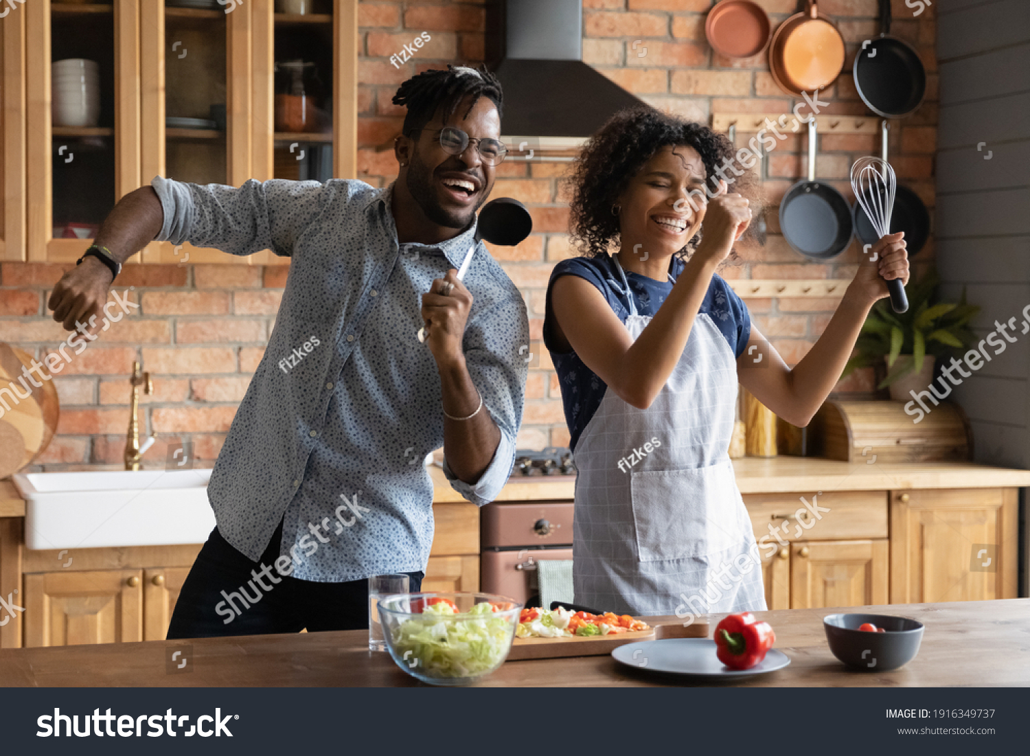 Close up overjoyed African American couple singing into kitchenware, having fun, dancing, listening to music in kitchen, cooking salad, excited wife and husband holding whisk and ladle a microphone #1916349737
