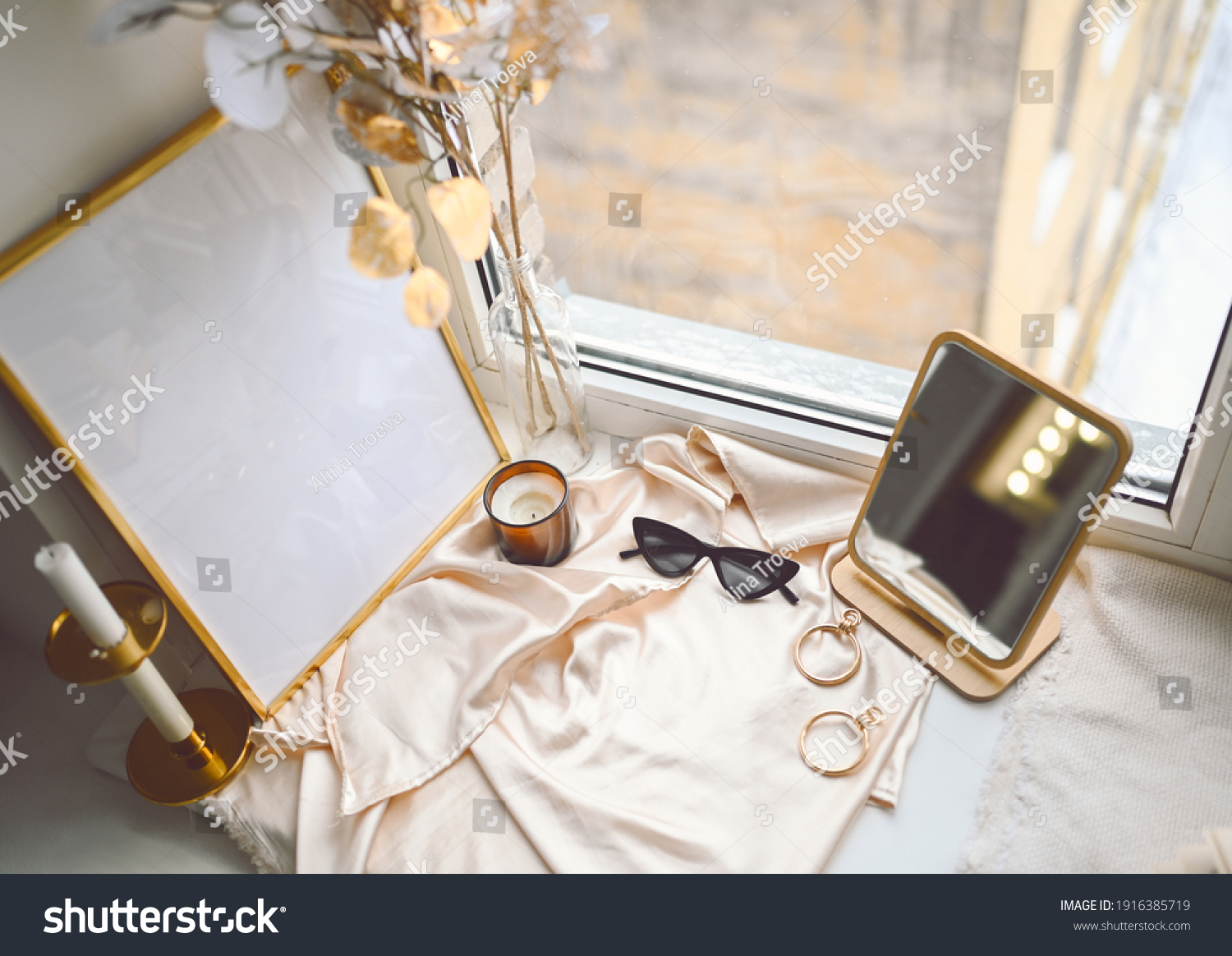 Cozy spring still life feminine scene golden shades. Female styled window sill minimalistic composition. Empty picture mock up poster frame, elegant accessories mirror earrings, dried flowers, candle #1916385719