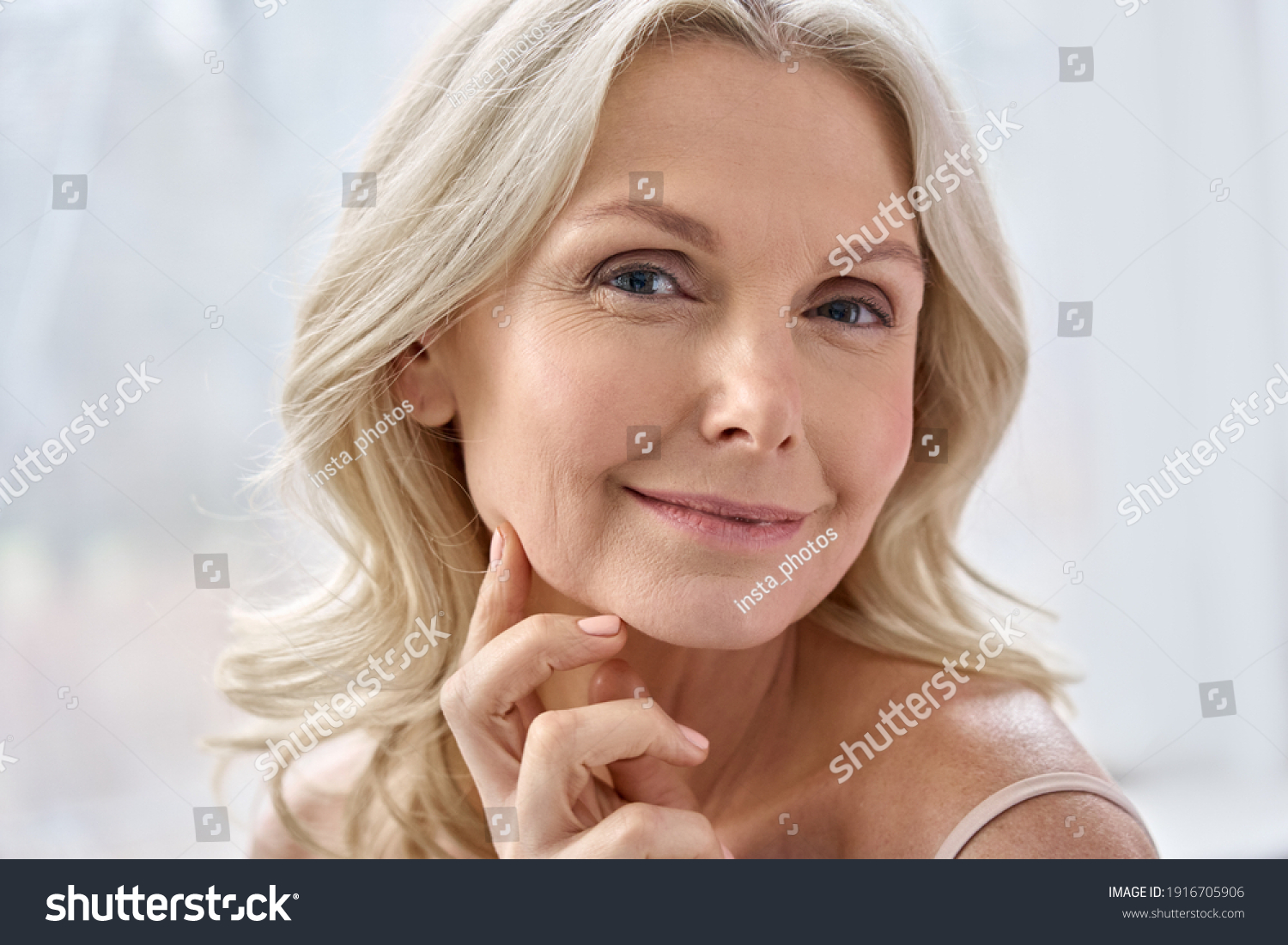 Smiling happy attractive 50s middle aged mature blond woman, old lady looking at camera advertising anti age face skin and body care treatment cosmetics posing in bathroom. Close up headshot portrait #1916705906