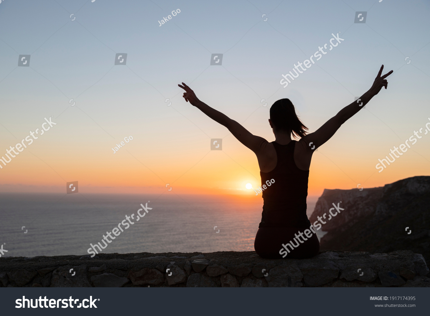 Backlit silhouette of a woman watching the sunrise over the sea making victory sign #1917174395