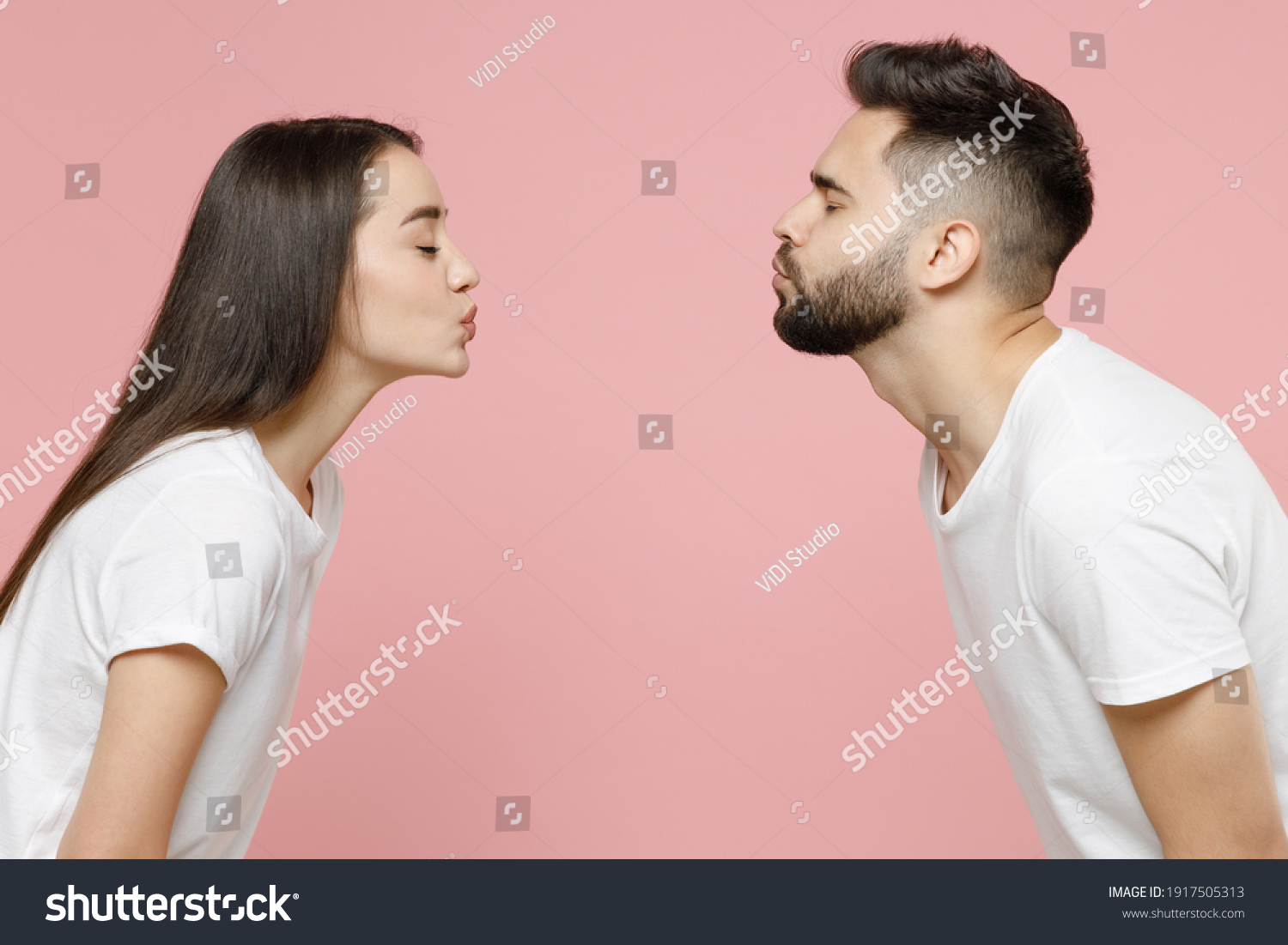 Side profile view young cheerful couple two friends man woman in white basic t-shirts kissing each other with closed eyes while standing face to face isolated on pastel pink background studio portrait #1917505313