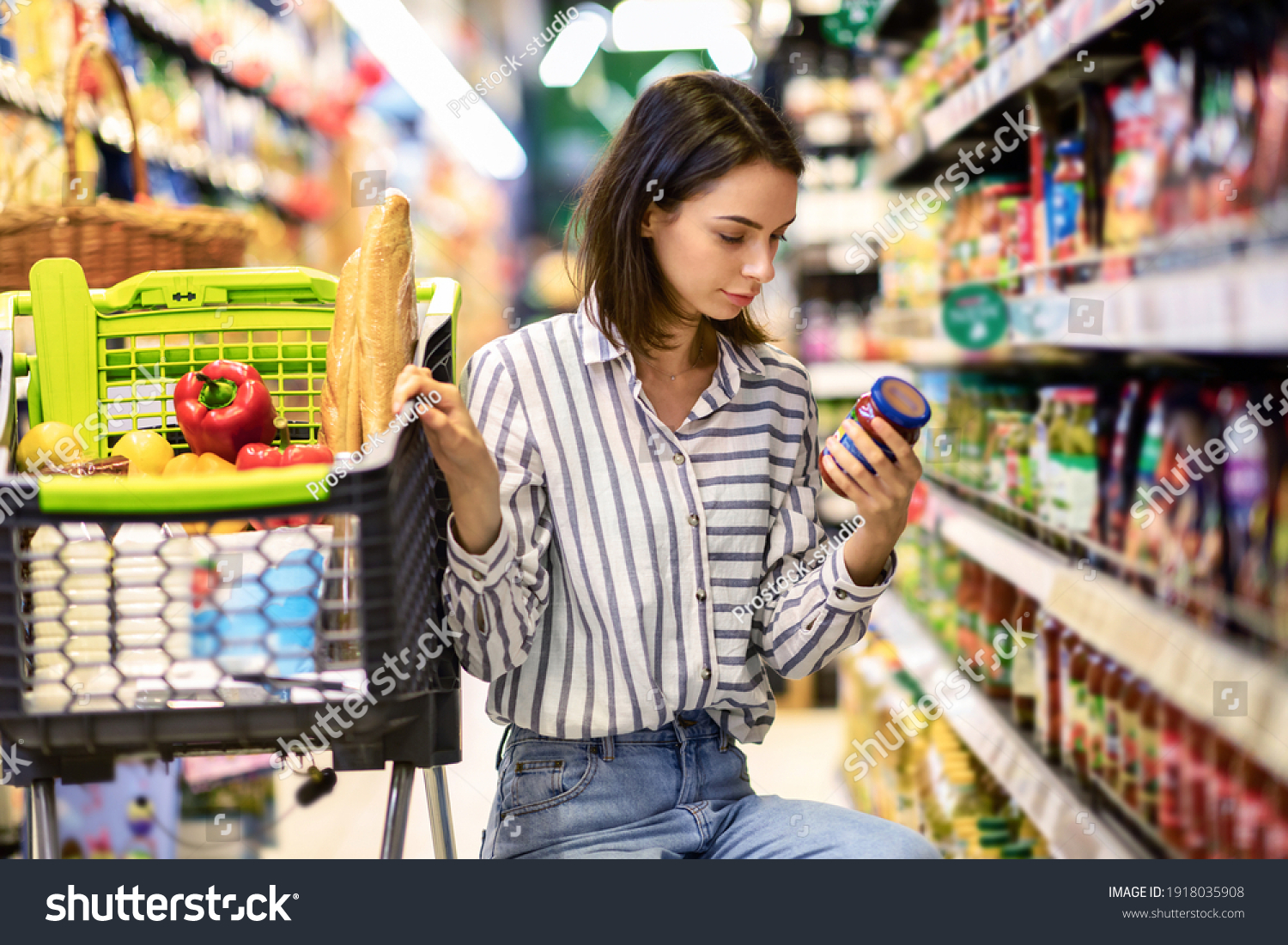 Consumption And Consumerism. Portrait Of Young Woman With Shopping Cart In Market Buying Groceries Food Taking Products From Shelves In Store, Holding Glass Jar Of Sauce, Checking Label Or Expiry Date #1918035908