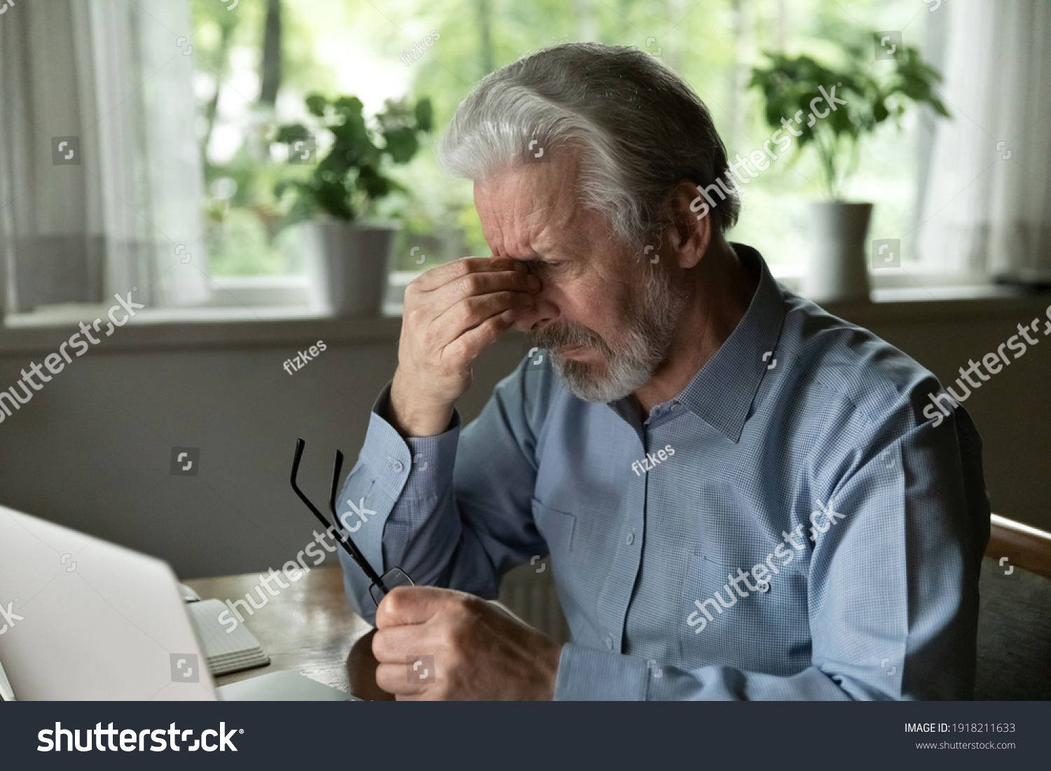 Close up stressed mature man massaging nose bridge, holding glasses, sitting at desk with laptop, elderly grey haired male suffering from eye strain, dry eyes syndrome after long laptop use #1918211633