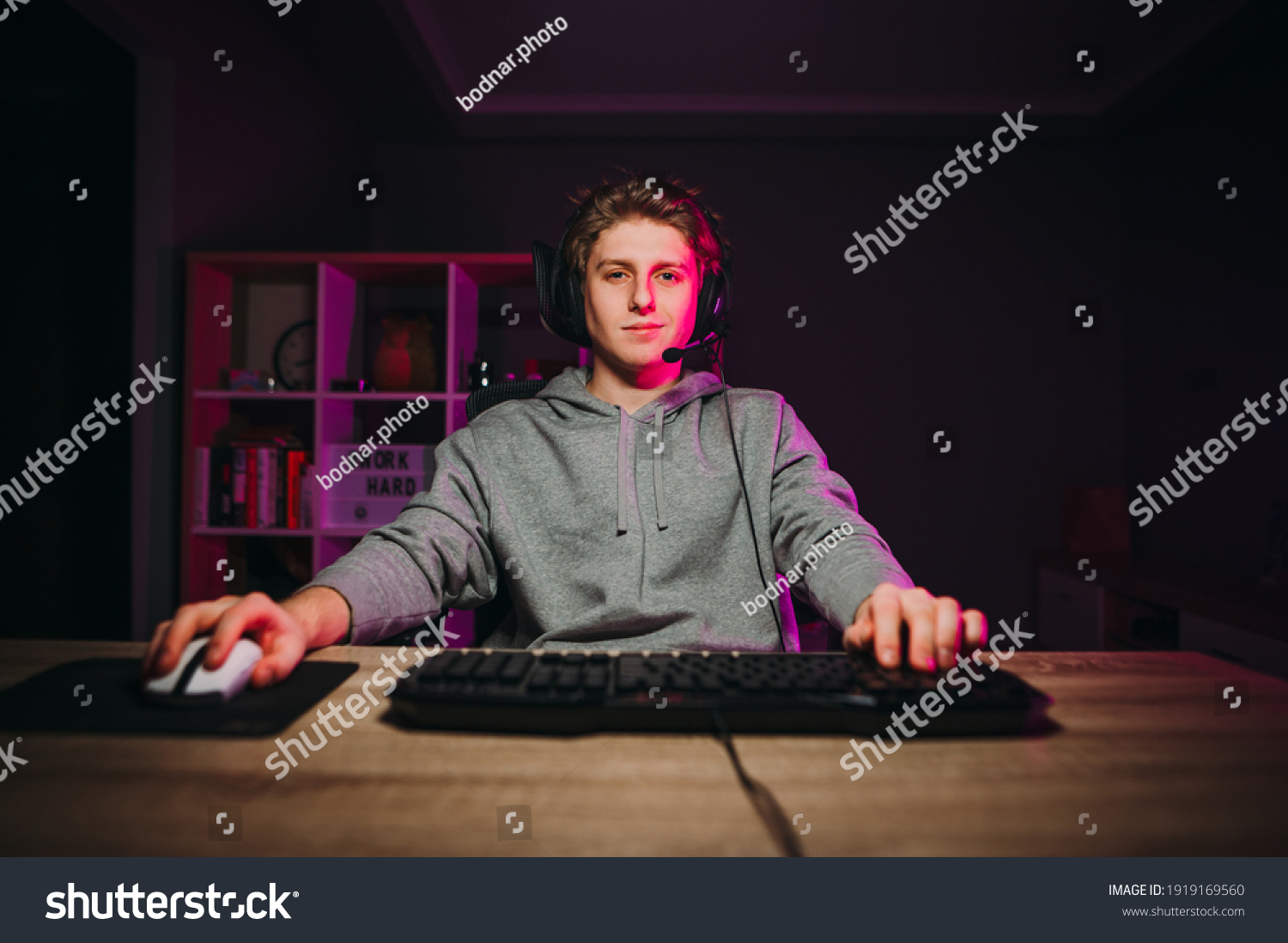 Handsome young man in casual clothes and headset sitting at a table in a room with purple lights and playing video games on the computer with a smile on his face looking at the screen. #1919169560