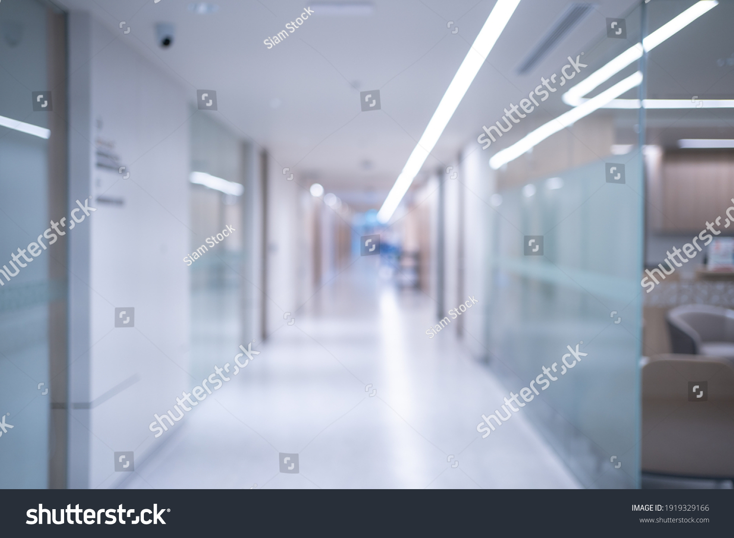 Blurred background of an interior of a modern hospital with an empty long corridor, there are treatment rooms and waiting room for patients and families between the corridor with bright white lights. #1919329166