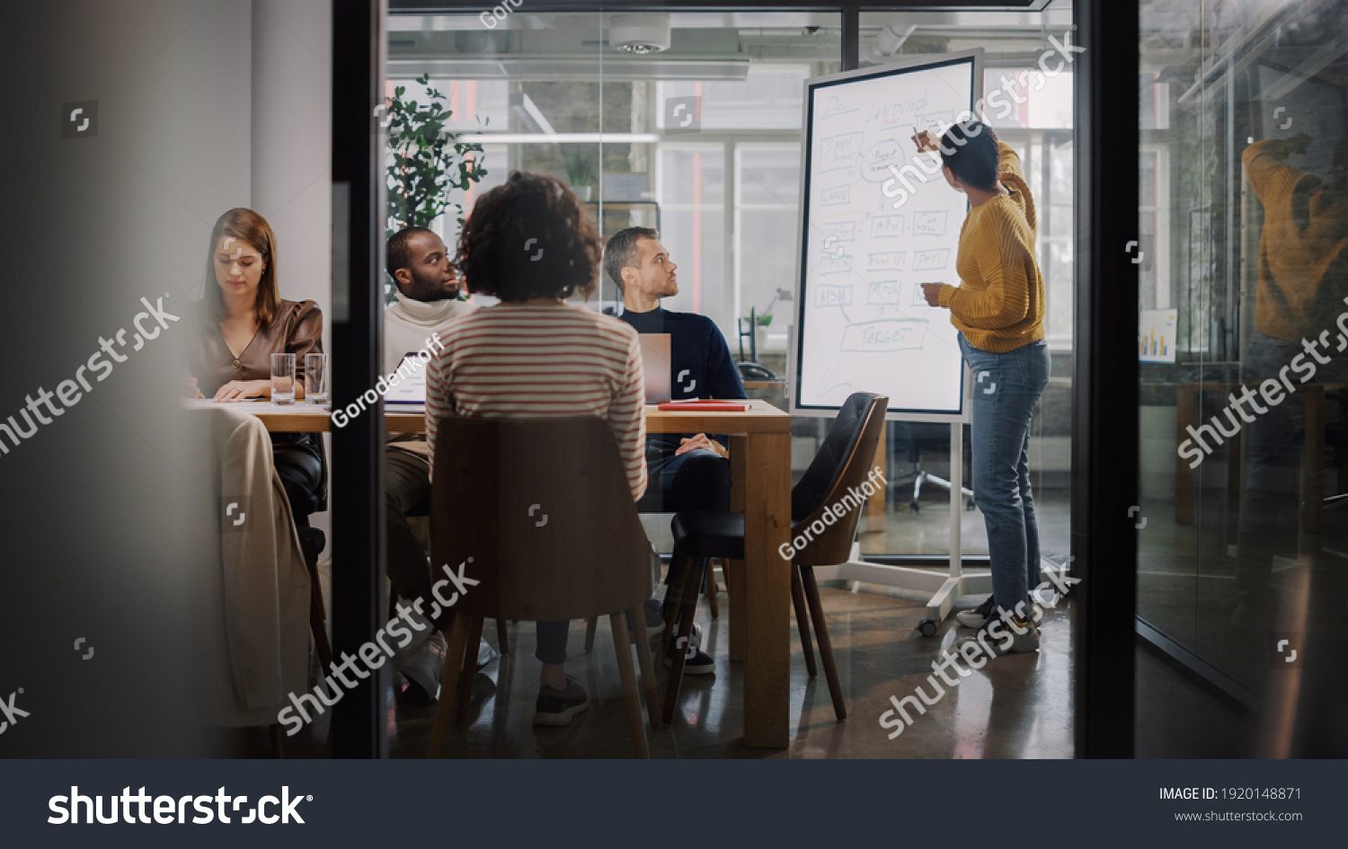 Project Manager Makes a Presentation for a Young Diverse Creative Team in Meeting Room in an Agency. Colleagues Sit Behind Conference Table and Discuss Business Development, User Interface and Design. #1920148871