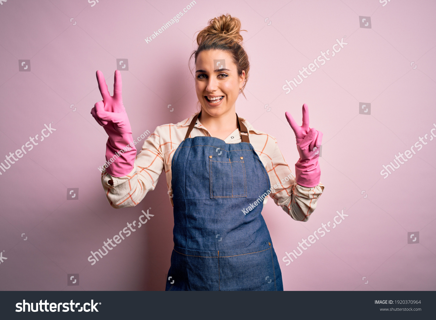 Young beautiful blonde cleaner woman doing housework wearing arpon and gloves smiling looking to the camera showing fingers doing victory sign. Number two. #1920370964