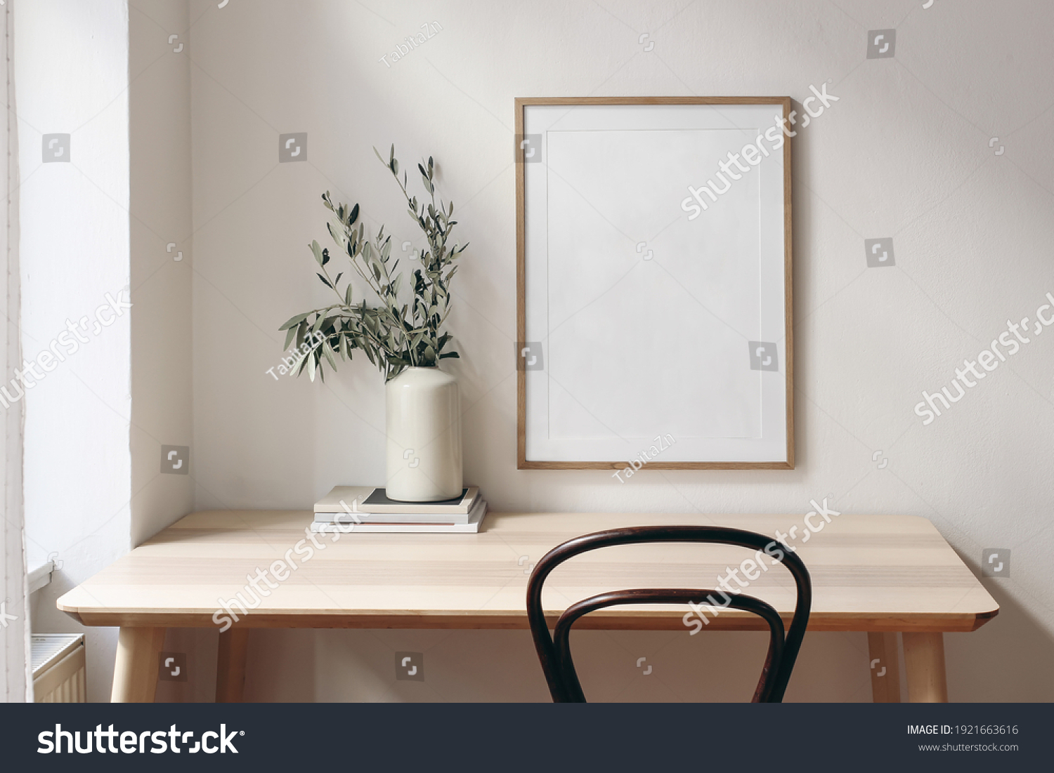 Home office concept. Old books, empty vertical wooden picture frame mockup hanging on white wall. Wooden desk, table. Vase with olive branches. Elegant working space. Scandinavian interior design. #1921663616