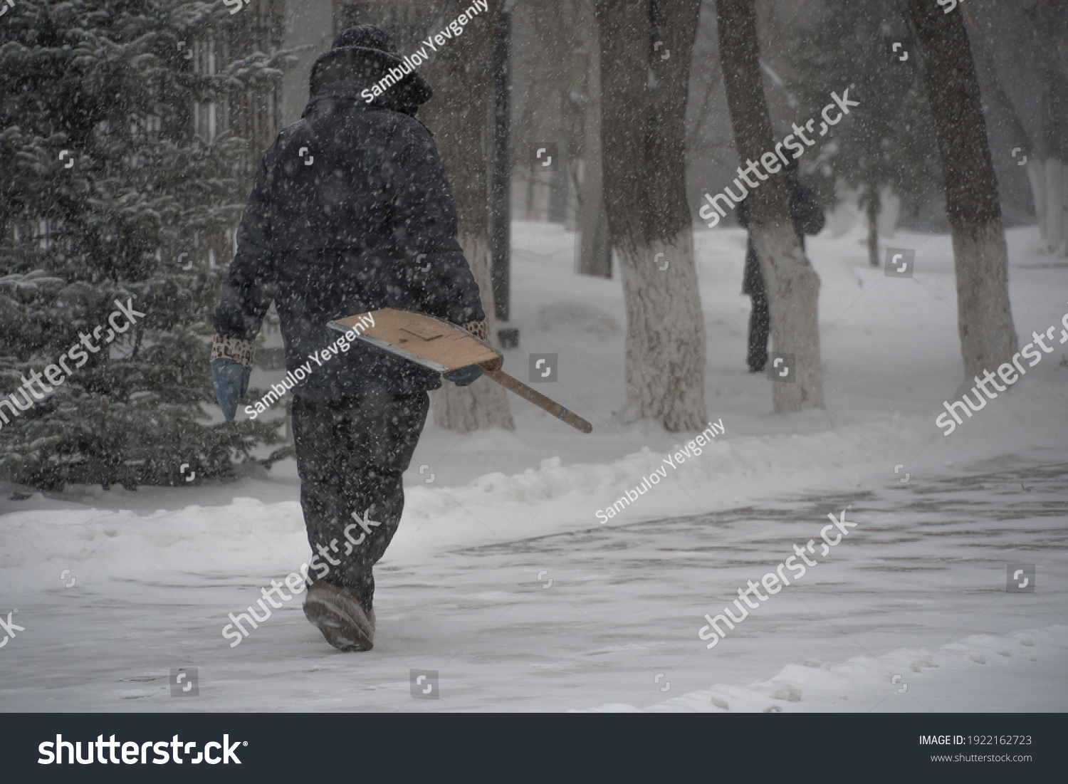 A working man of the municipal service with a snow shovel walks along the road in a storm, blizzard or snowfall in winter in bad weather in the city.Extreme winter weather conditions in the north. #1922162723