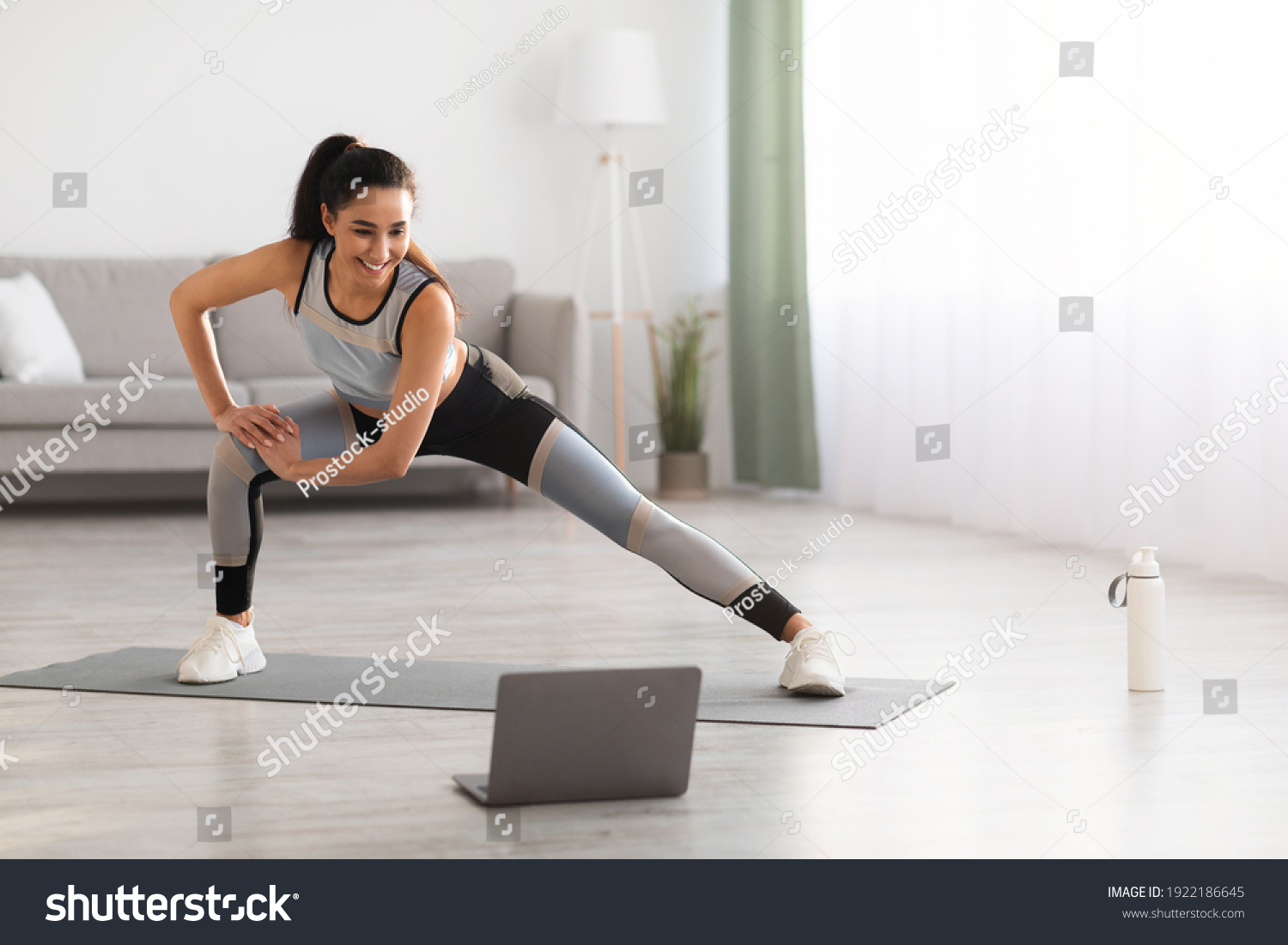 Sporty young brunette woman exercising at home, stratching her legs, using laptop, watching fitness video on Internet or having online fitness class, living room interior, copy space #1922186645