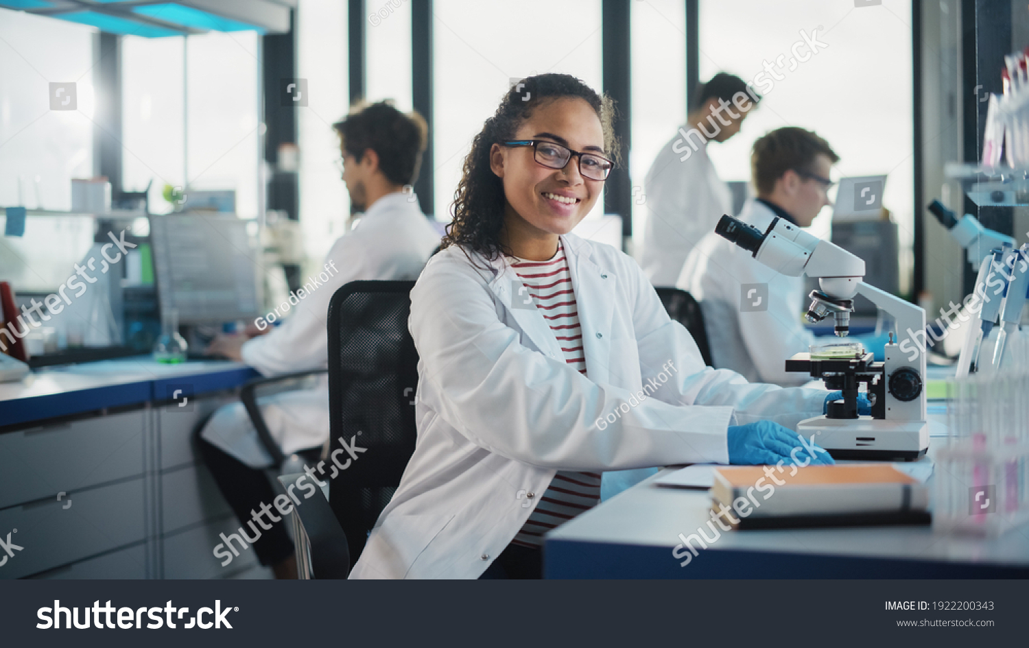 Medical Science Laboratory: Beautiful Black Scientist is Using Microscope, Looking at Camera and Smiling Charmingly. Young Biotechnology Science Specialist, Using Technologically Advanced Equipment. #1922200343