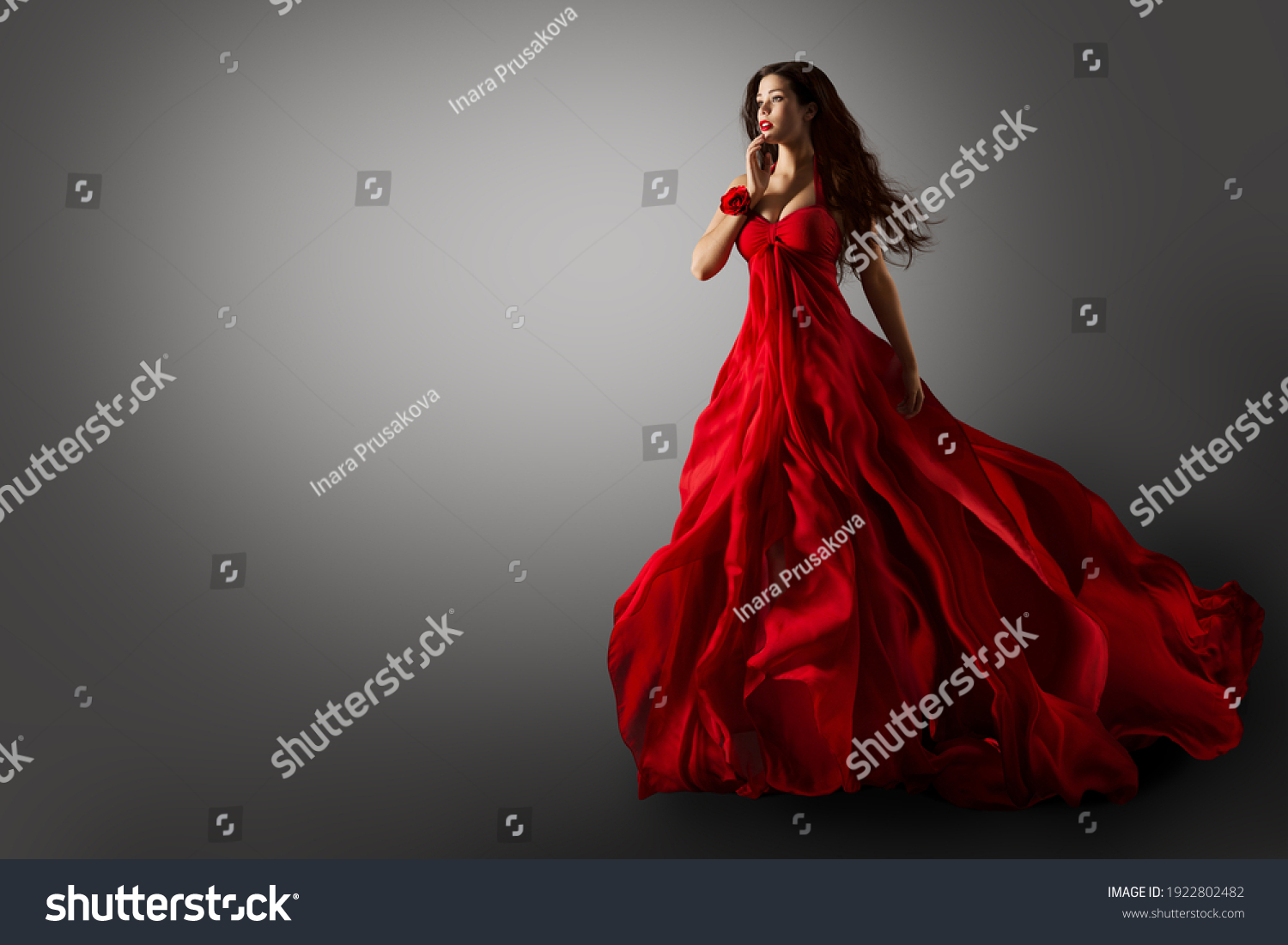 Fashion Woman in Red Dress. Beauty Model dancing in Long Evening Gown fluttering on Wind Black Hair flying in Air over Gray Background #1922802482