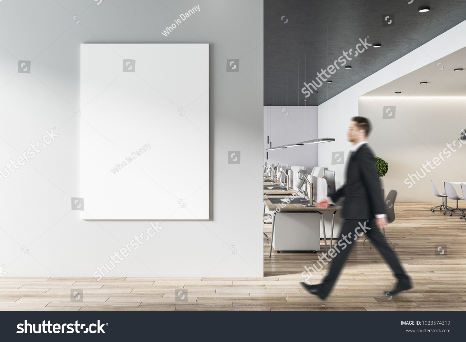 Businessman goes to blank white poster on light grey wall in modern open space office with light furniture and wooden floor. Mock up #1923574319