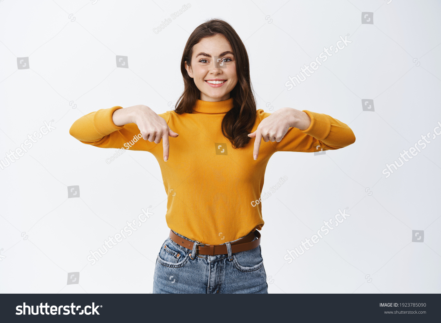 Smiling confident girl pointing fingers down to show advertisement, showing promo offer on bottom empty space, standing against white background. #1923785090