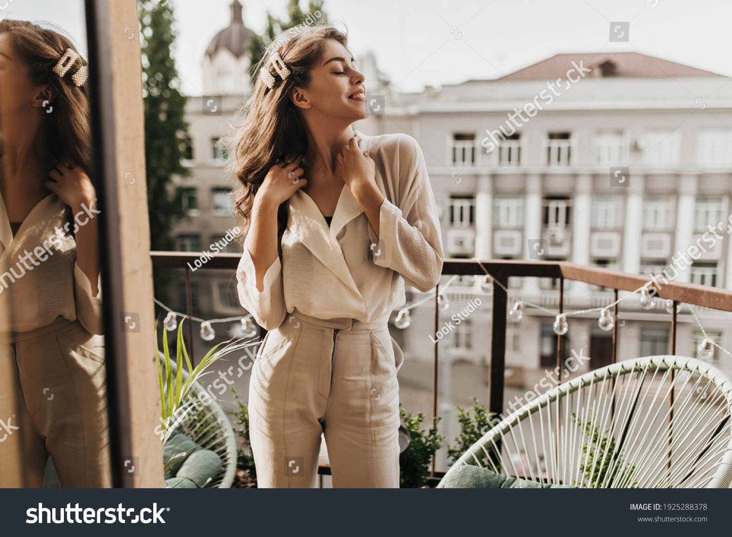 Curly woman enjoys sunny day on terrace. Pretty dark-haired lady in beige blouse and stylish pants poses on balcony #1925288378