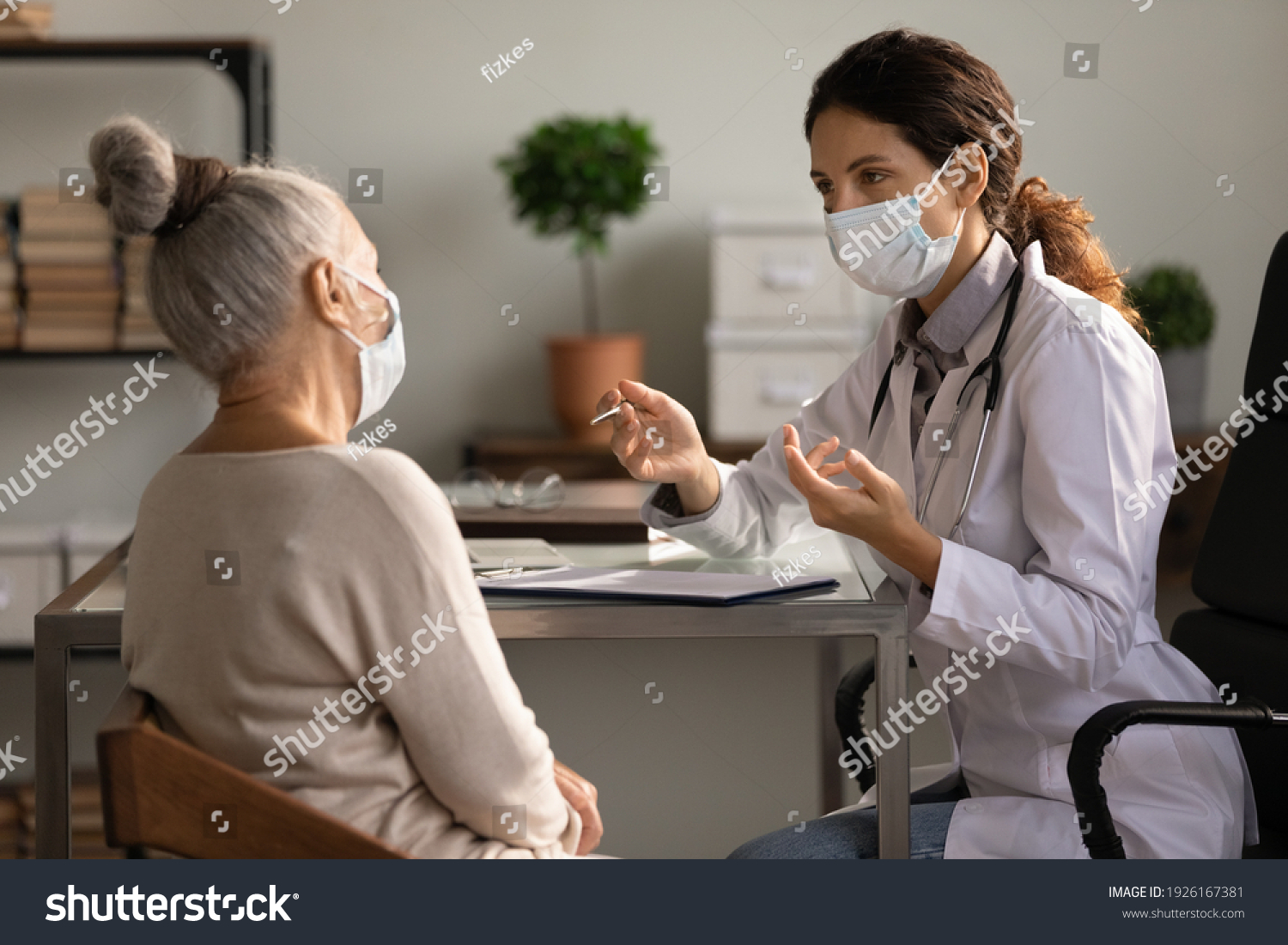 Close up serious doctor wearing medical face mask consulting mature woman patient at appointment in office, physician explaining treatment, giving recommendations, elderly generation healthcare #1926167381