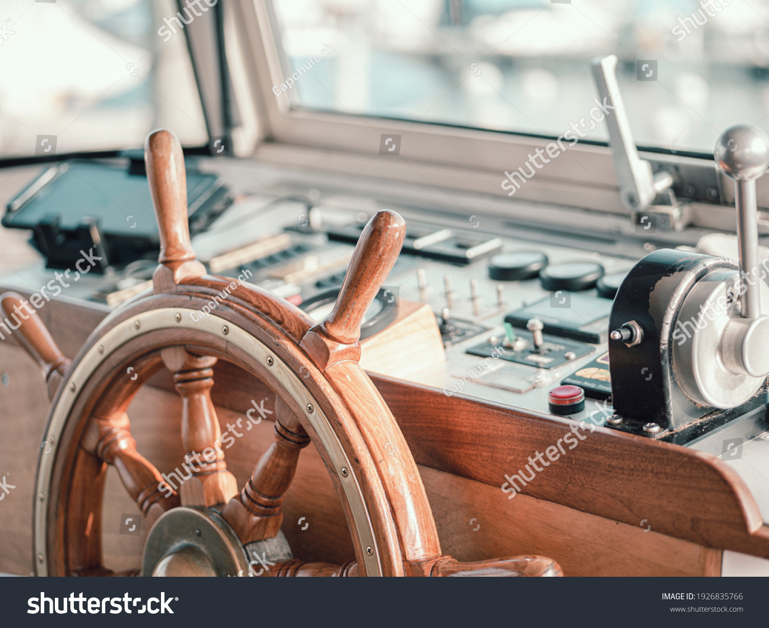 Close up of the interior of a large motor boat. The steering wheel and motor controls can be seen and the boat harbor slurred through the glass boat window. #1926835766