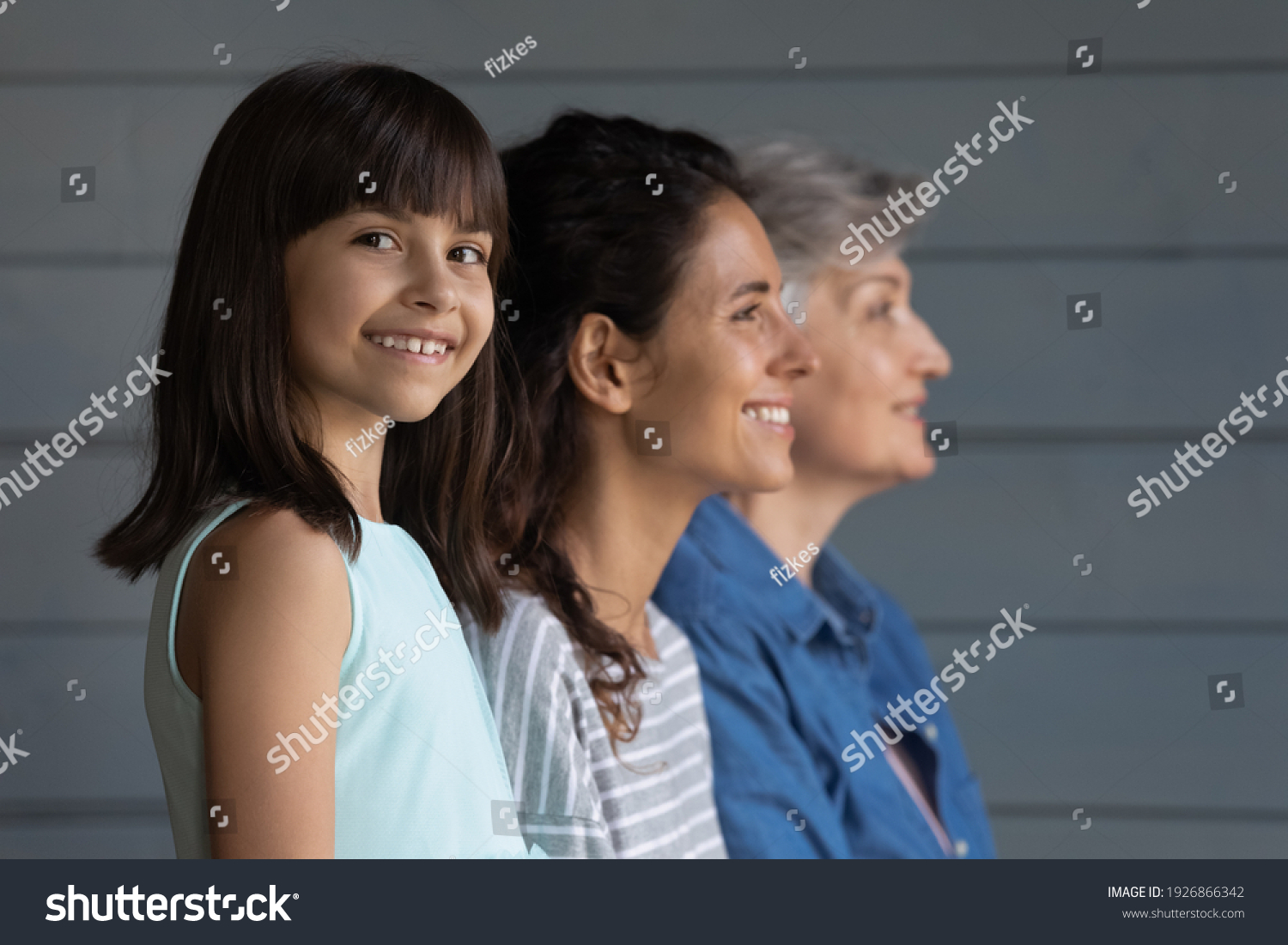 Portrait of happy little Hispanic 7s girl child look at camera smiling, mom and grandmother on background look in distance. Growing generation or small Latino kid with mother and senior grandparent. #1926866342