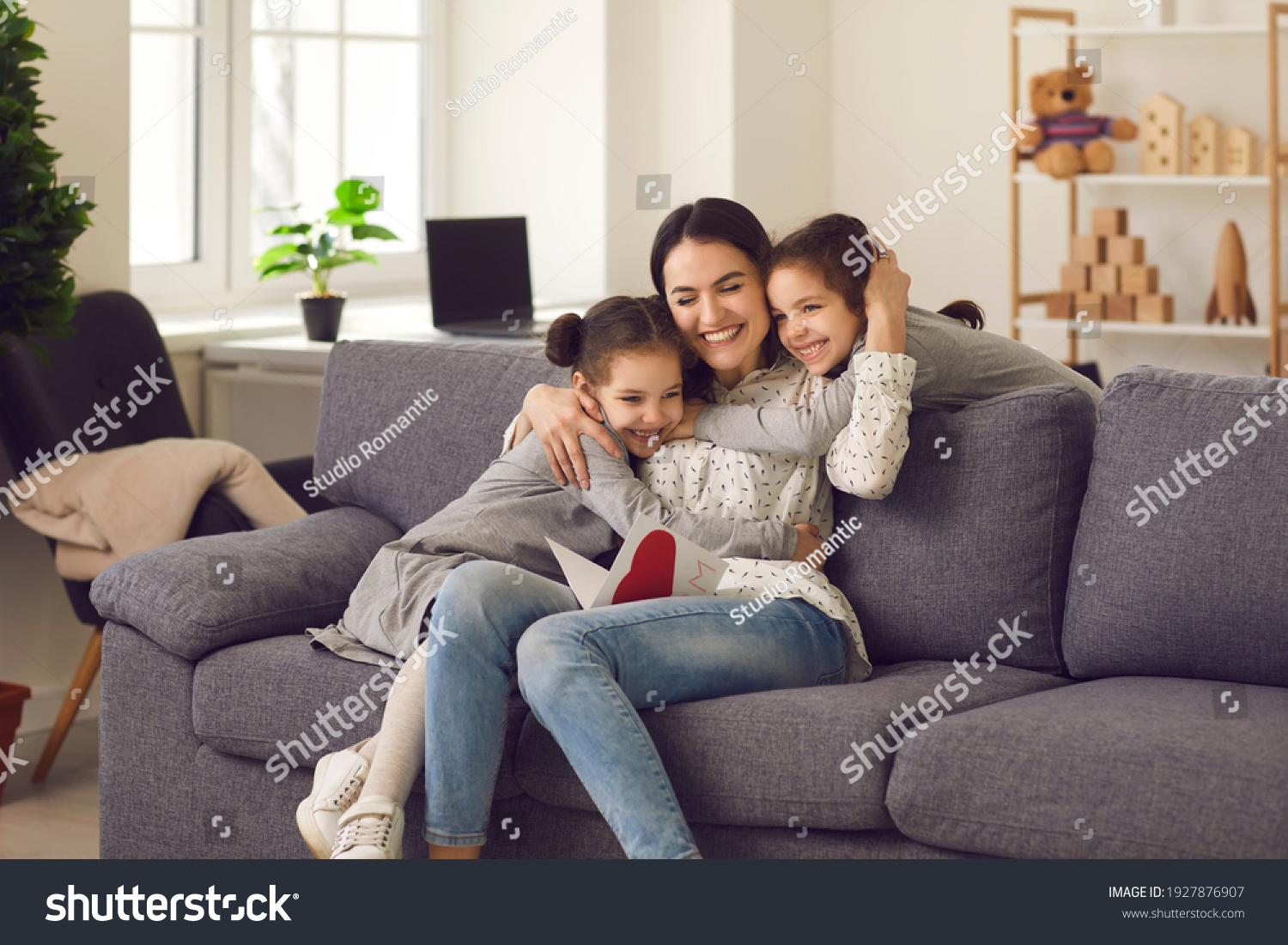 Happy loving mother hugs her daughters, who gave her a greeting card for the holiday. Sisters made a handmade card with a heart and congratulated the mother on Mother's Day, birthday or Women's Day. #1927876907