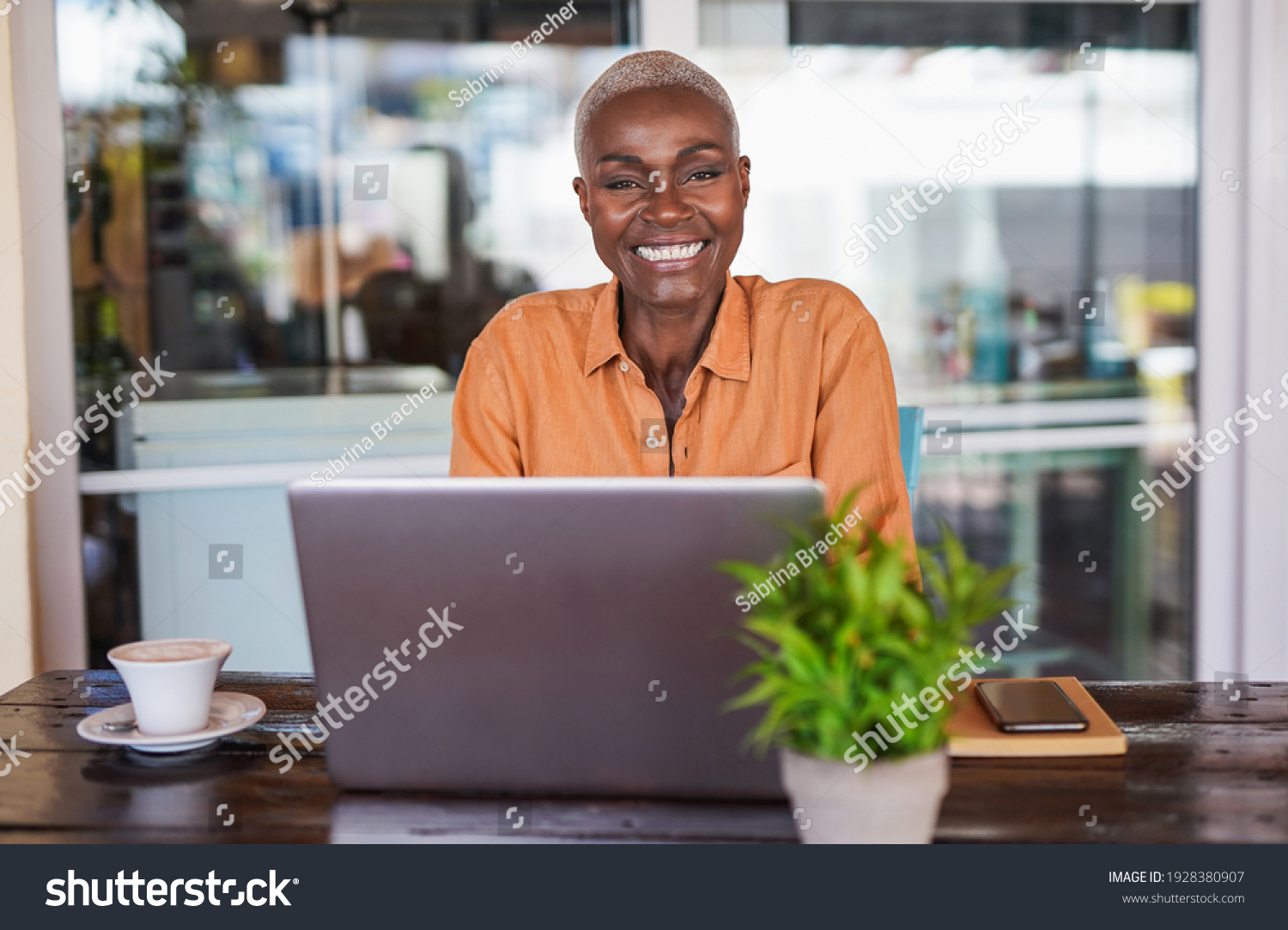 Beautiful mature african woman sitting in a bar outdoor and working with laptop - Black woman looking in camera and smiling #1928380907