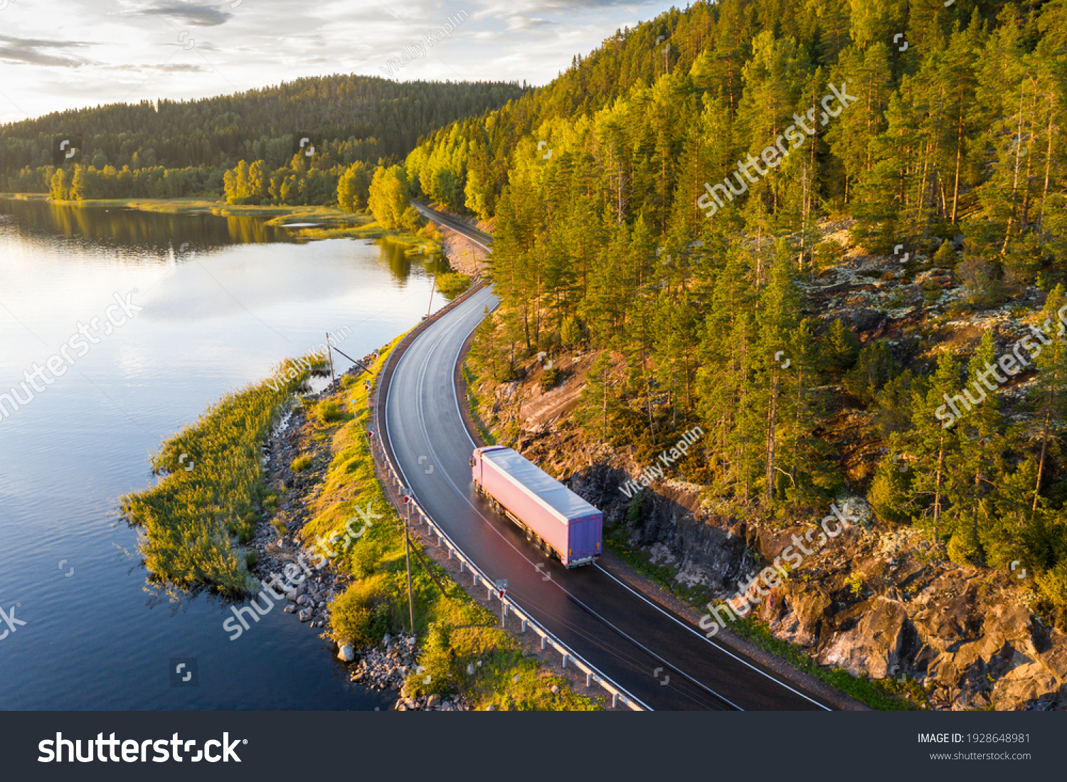 Aerial view of semi truck with cargo trailer on road curve at lake shore with green pine forest. Transportation background. Beautiful nature landscape at sunset in Republic of Karelia, Russia #1928648981