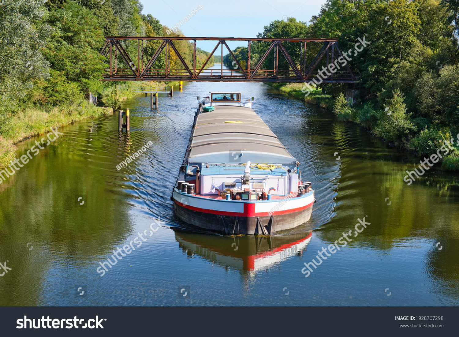 a cargo canal ship passes under an old iron bridge on a sunny autumn day, reflections are to be seen in the water, canal system in the outskirts of Berlin Germany #1928767298