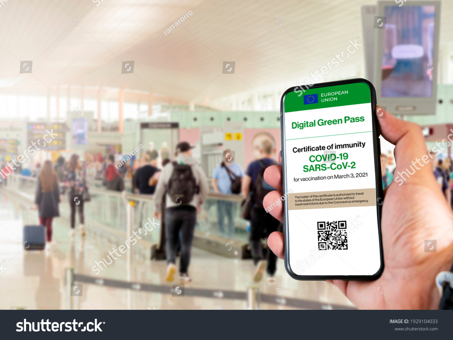The digital green pass of the european union with the QR code on the screen of a mobile held by a hand with blurred airport in the background. Immunity from Covid-19. Travel without restrictions. #1929104033